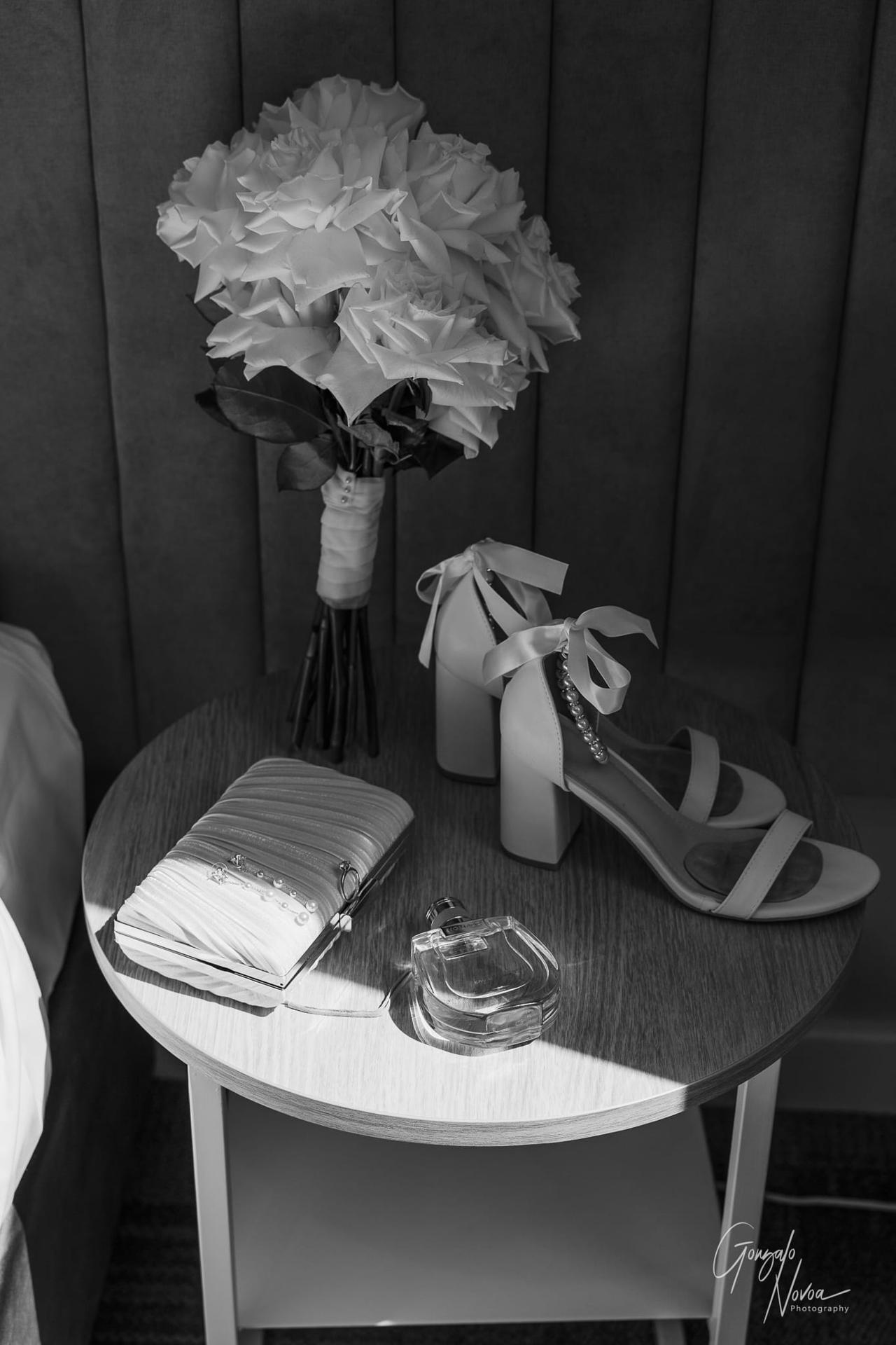 Bouquet of white roses, heeled shoes with ribbons, a pearl clutch, and a perfume bottle on a wooden table. Soft light and elegant mood.