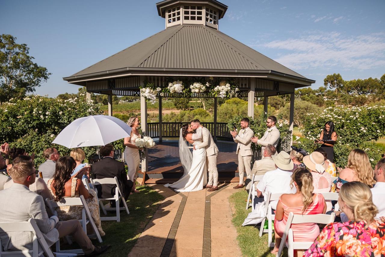 Bride and groom kiss under a gazebo adorned with flowers as guests seated outdoors applaud. A sunny, joyful wedding scene.
