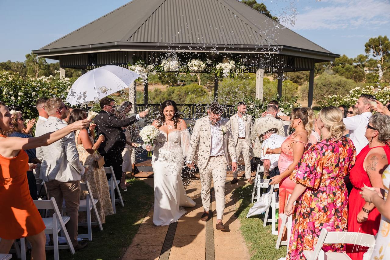 Bride and groom walk down the aisle under confetti, surrounded by cheering guests in colorful outfits. Outdoor gazebo setting.