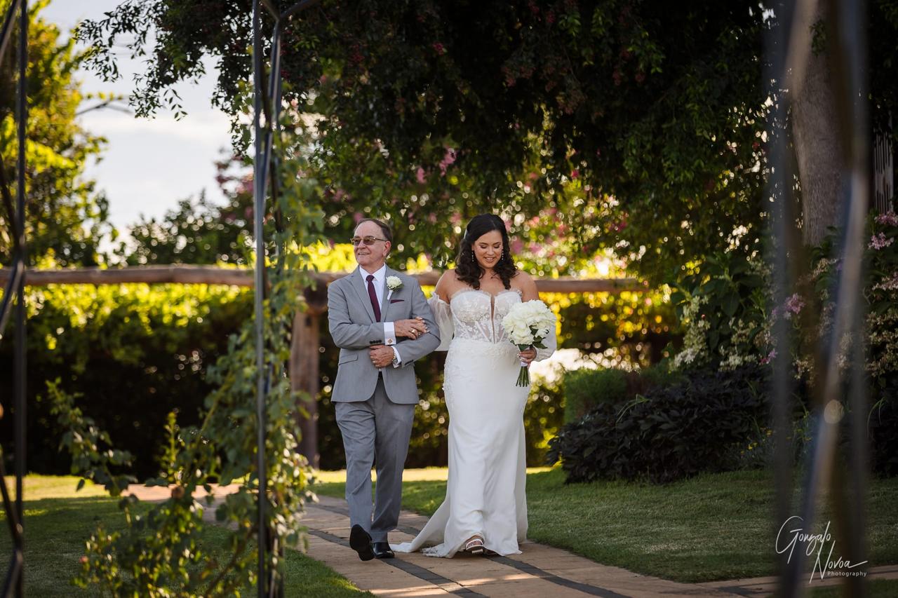 A bride in a white dress holds a bouquet, walking arm-in-arm with a man in a gray suit. They smile in a lush garden setting.