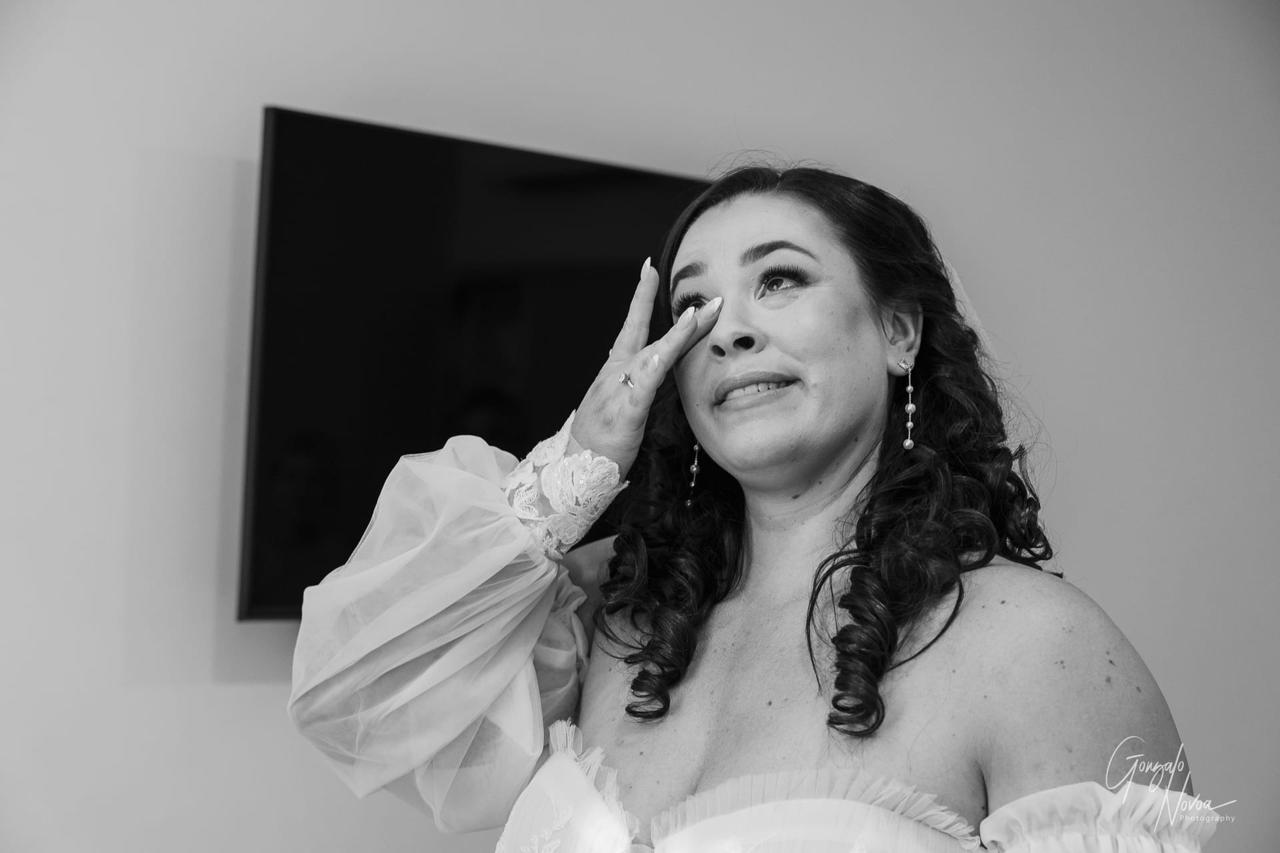 Bride in lace dress wipes tears, smiling with emotion. Black and white image with blurred background, conveying joy and sentiment.