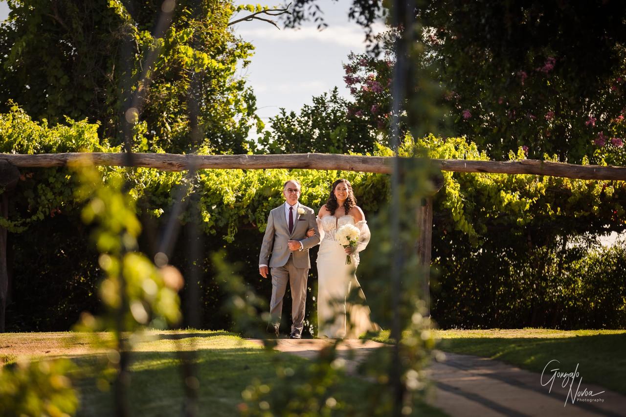 Bride in white dress walks arm-in-arm with an elderly man in a suit under a wooden archway with greenery. Sunlight filters through trees.