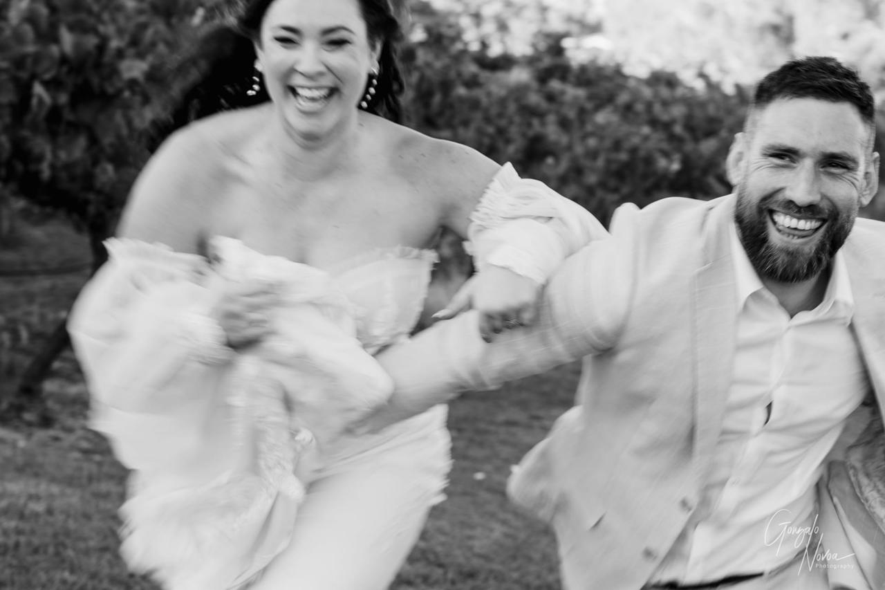 A joyful couple runs hand-in-hand in a garden. The woman wears a flowing dress, the man a light suit. Both are smiling, blurred motion.