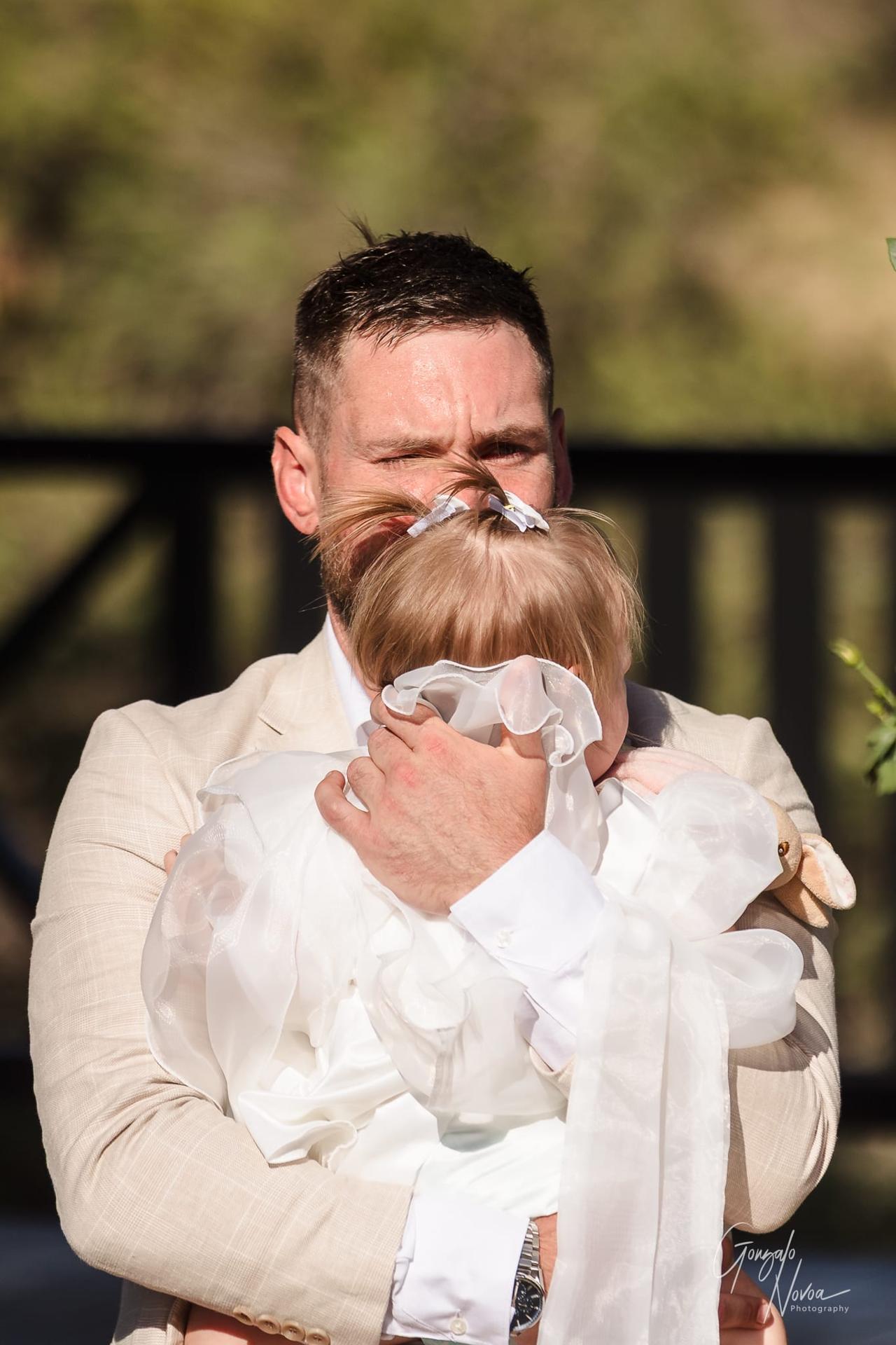 A man in a beige suit embraces a child in a white dress outdoors. The child has a small hair bow. The mood is tender and serene.