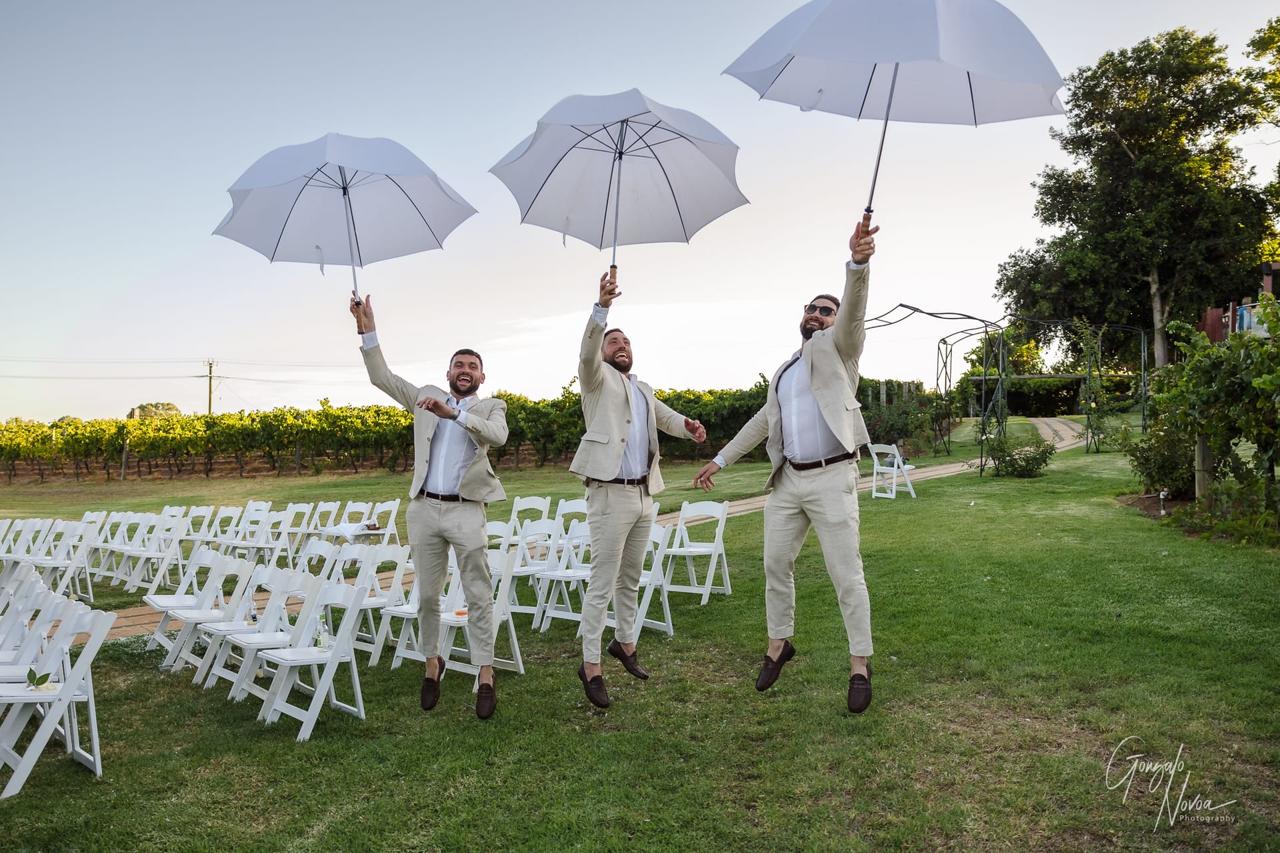 Three men in beige suits joyfully jump while holding white umbrellas in a garden setting with empty white chairs and a vineyard background.