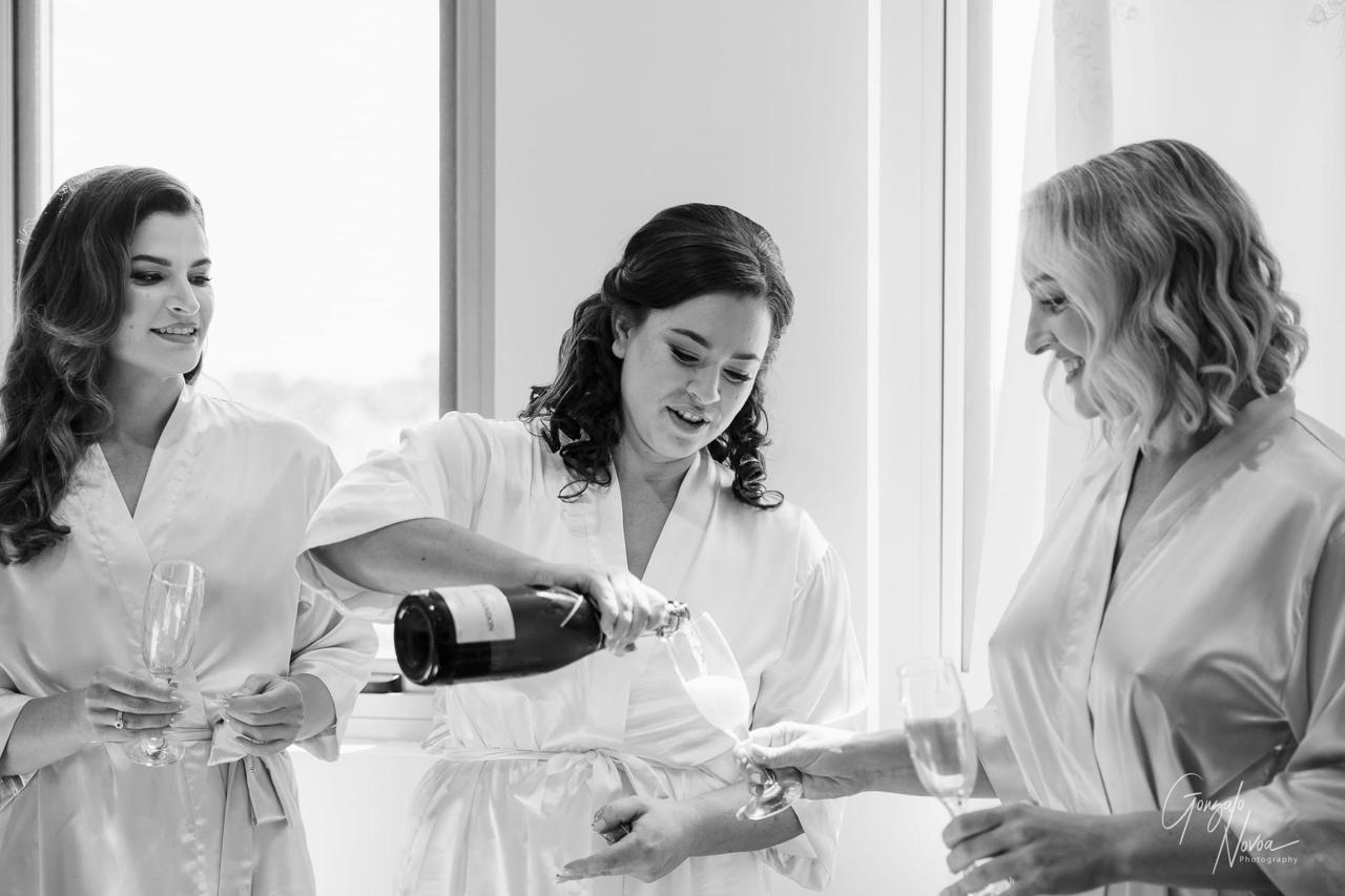 Three women in silk robes, smiling, as one pours champagne into glasses by a window. Bright light and joyful mood.