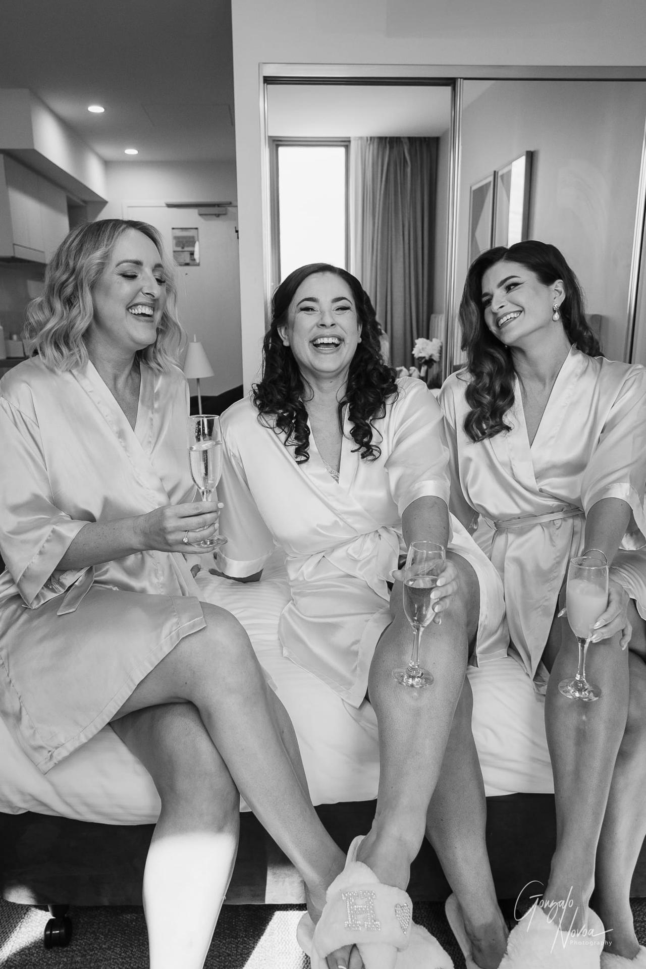 Three women in white robes laugh and hold champagne glasses, sitting on a bed in a bright room. The mood is joyful and celebratory.
