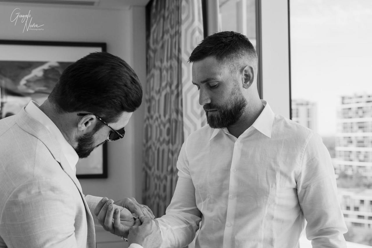Two men in a room, one in sunglasses adjusting the other's cufflink. They wear white shirts, with buildings visible through a window.