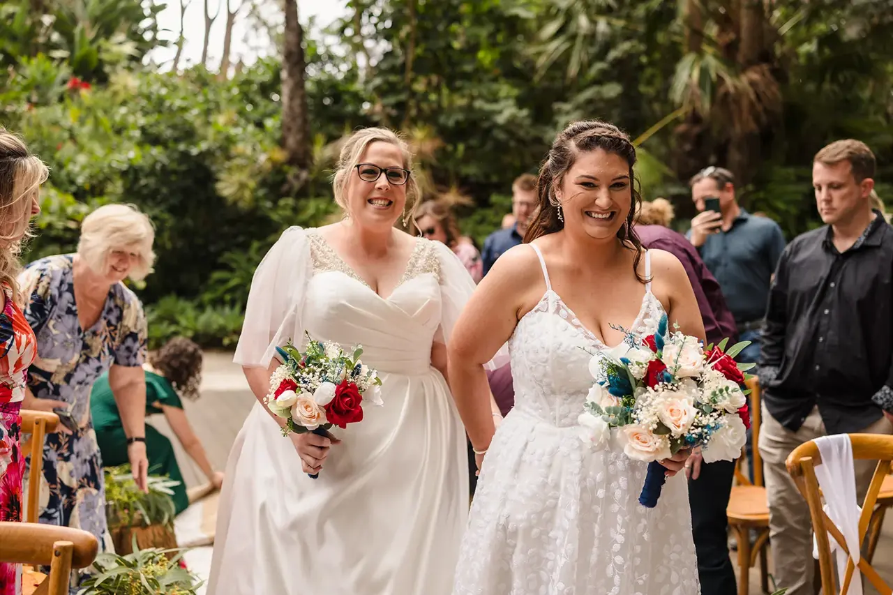 Kat and Jess walk down the aisle together side by side with their bouquets at their Wanneroo Botanic Gardens garden wedding ceremony