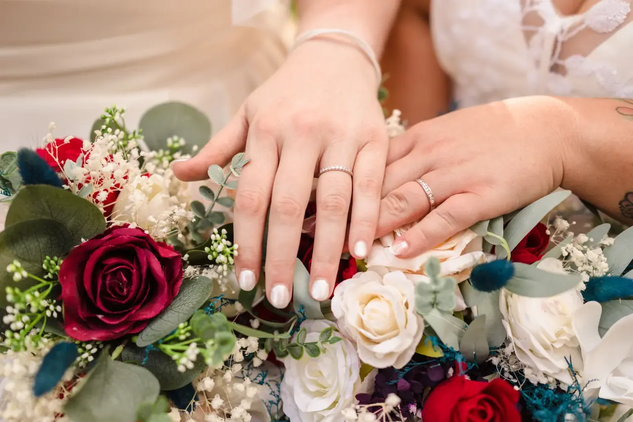 Kat and Jess's wedding rings on their hands, resting on their red rose and white bouquets