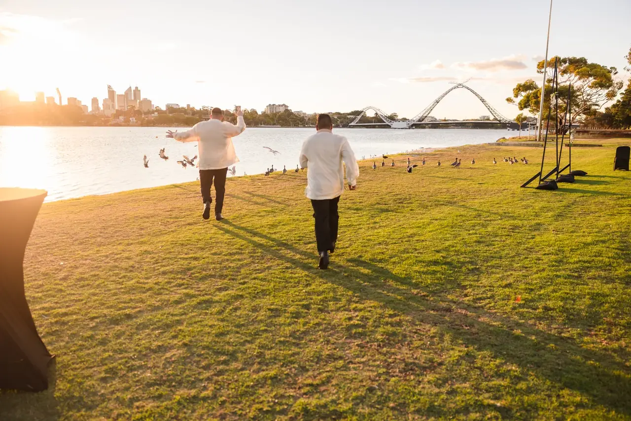 Lloyd and Mitch running along the riverside lawn at golden hour with the Perth skyline behind at Burswood on Swan