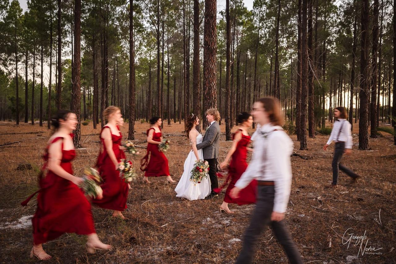Bride and groom stand in a forest, surrounded by blurred bridesmaids in red dresses and groomsmen in shirts and suspenders. Romantic setting.