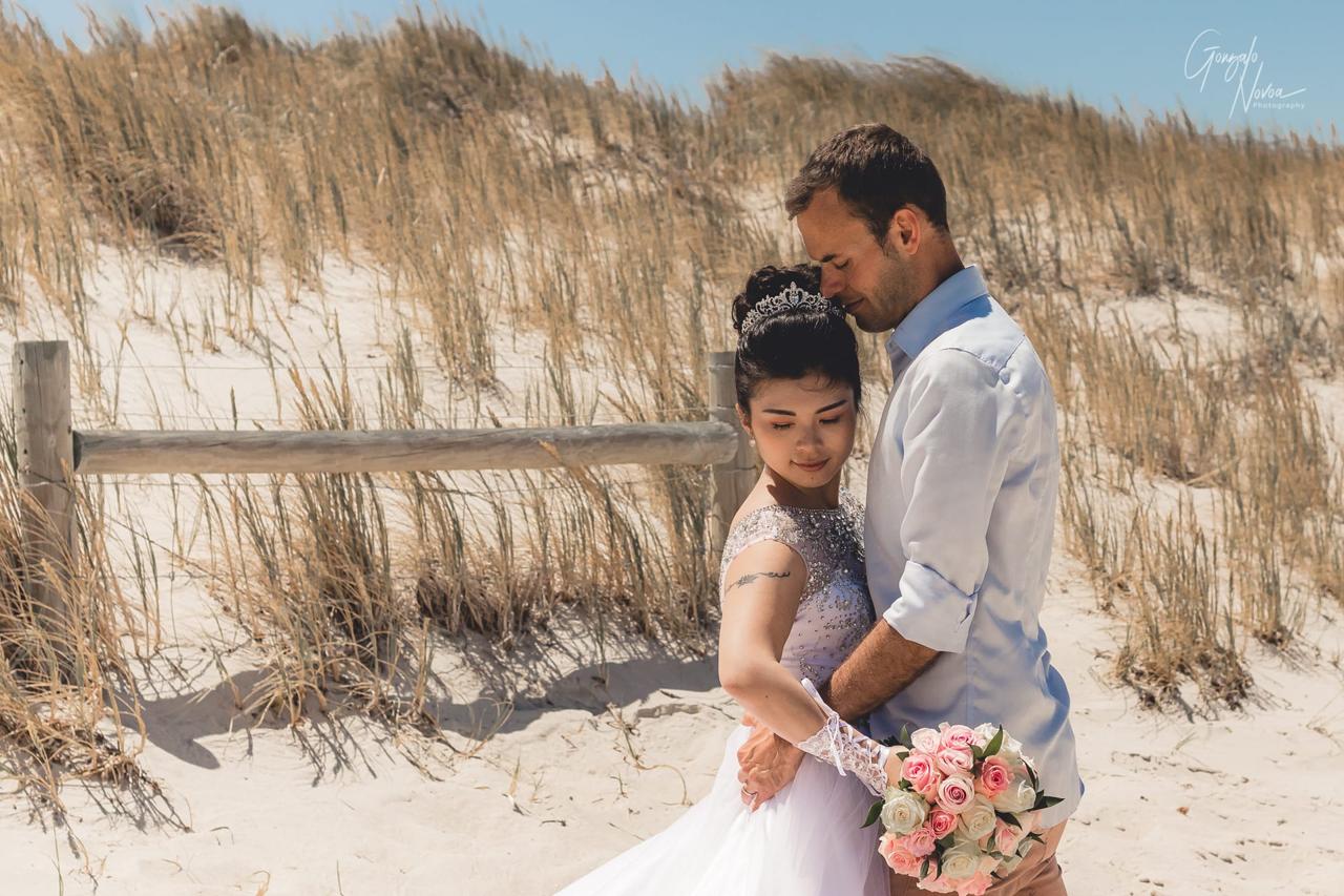 Bride and groom share a quiet, intimate moment during their First Look on a sandy beach, with the bride holding a bouquet of pink and white roses.