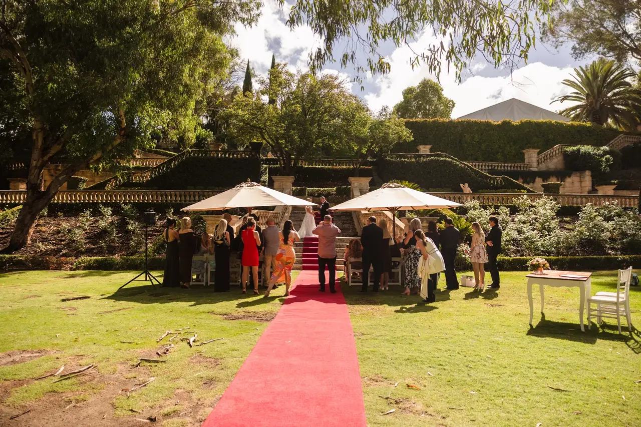 A ceremony on the lawn at Caversham House, guests under sunshades either side of a red carpet with the stone terraces behind