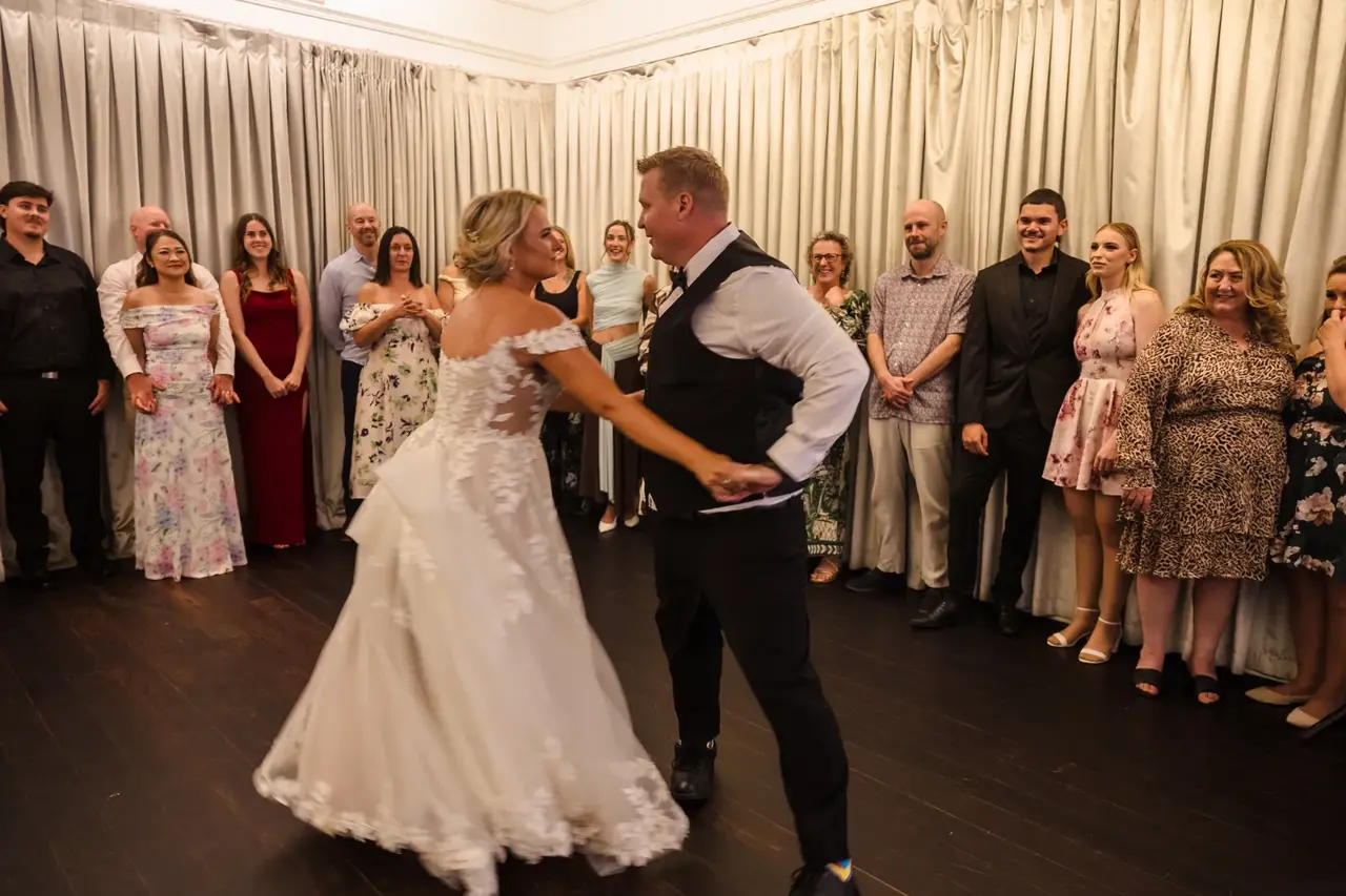 Rosie and Adam dance in a white reception room with guests gathered around them at Caversham House