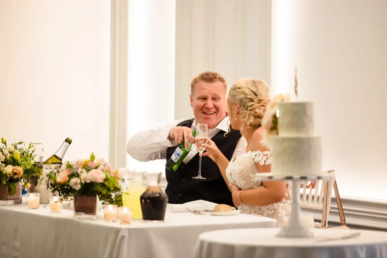 Rosie and Adam at the reception table at Caversham House, a bottle being poured beside the cake