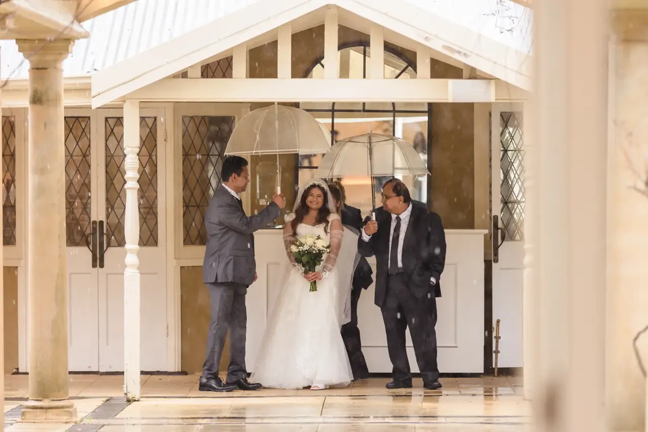 Naila stands at the garden house entrance between two men holding clear umbrellas