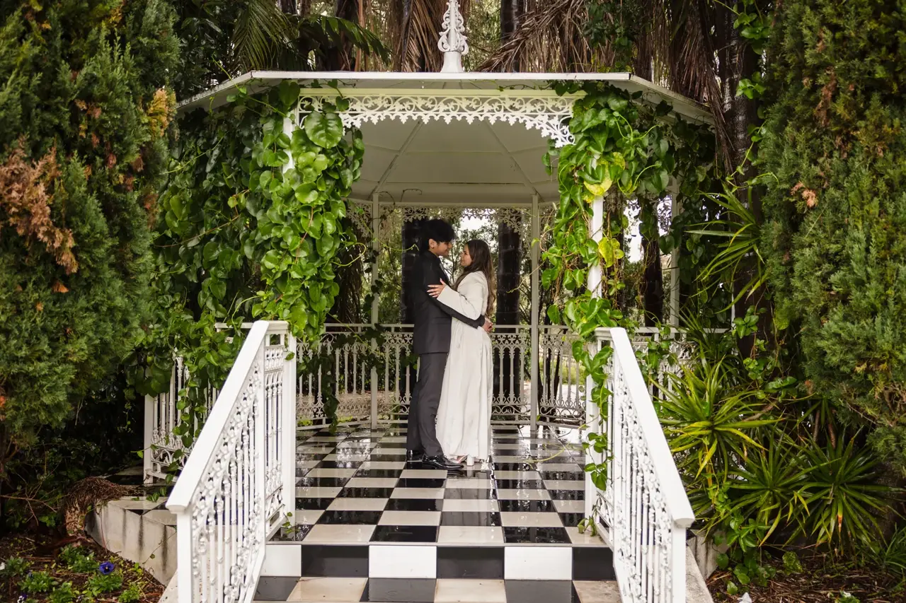 Naila and Kusal embrace on the black and white checkerboard steps of a white garden gazebo