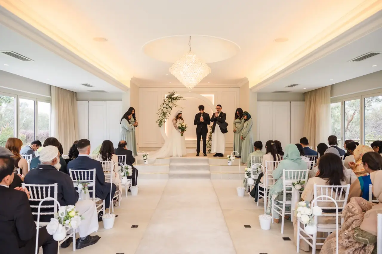 Guests sit on white chairs in the light-filled Garden House as Naila reaches Kusal at the floral arch