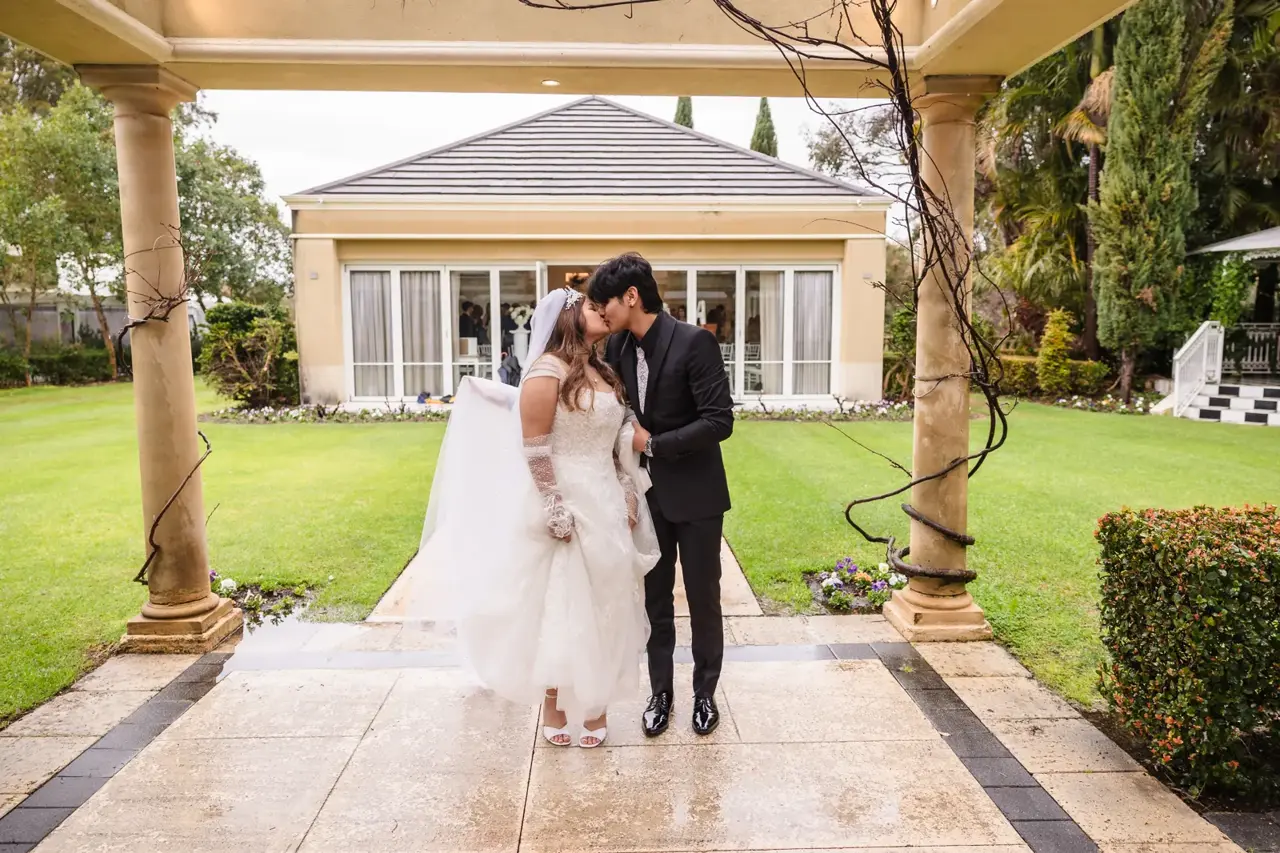 Naila and Kusal kiss between stone columns on a wet path, the Garden House behind them