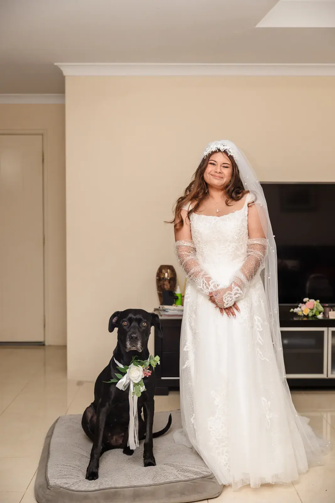 Naila in her wedding dress and veil stands beside her dog Luna, who wears a ribbon and greenery on her collar