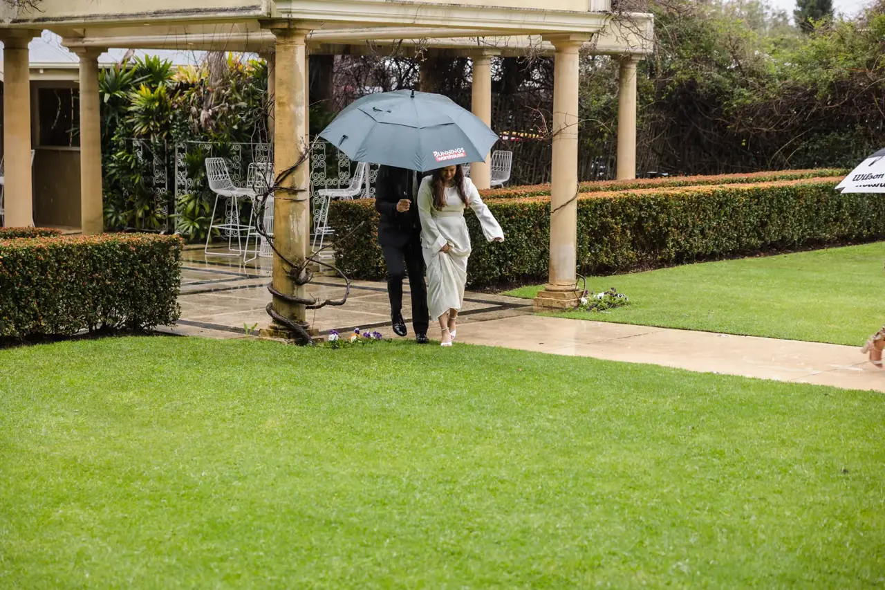 Naila and Kusal walk back across the lawn under one shared umbrella