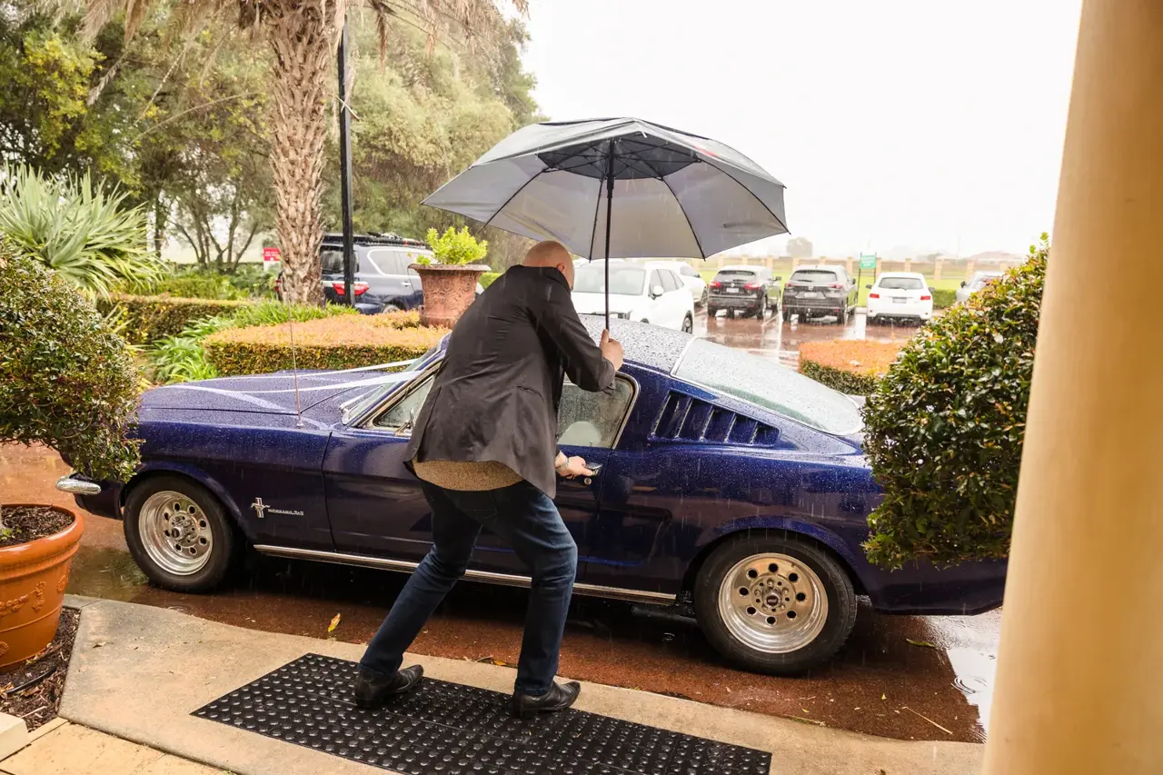 A man with an umbrella reaches for the door of a deep blue vintage Mustang in the rain