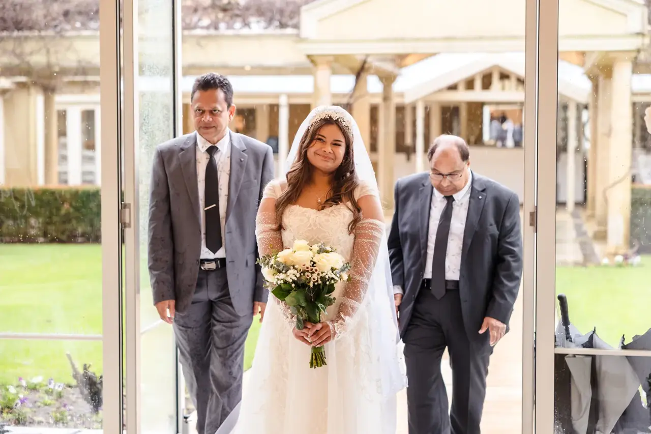 Naila walks in through the open glass doors between two men, rain falling on the garden behind
