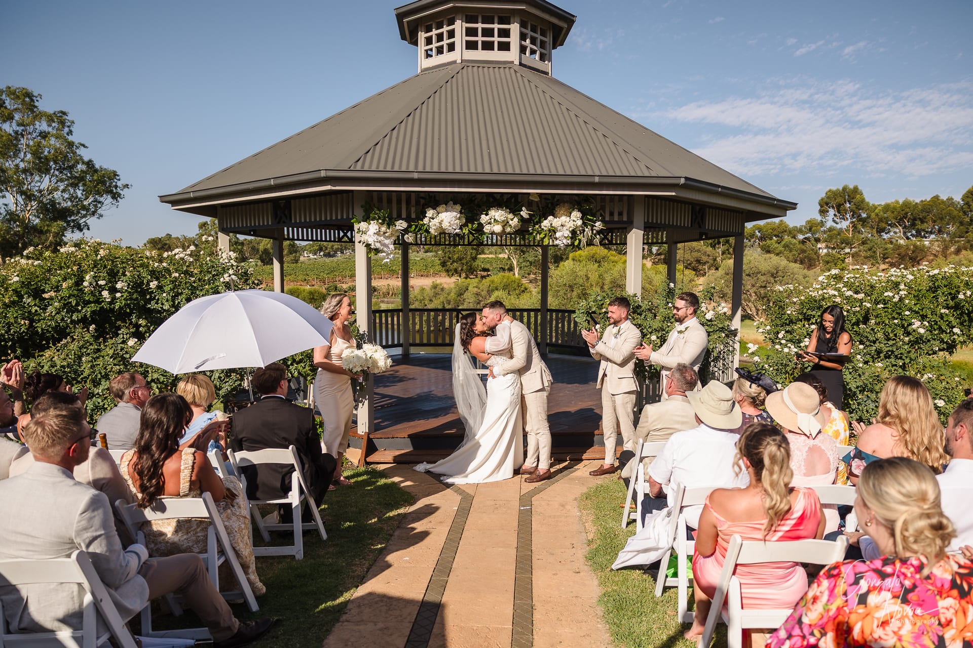 Bride and groom kiss under a gazebo adorned with flowers as guests seated outdoors applaud. A sunny, joyful wedding scene.