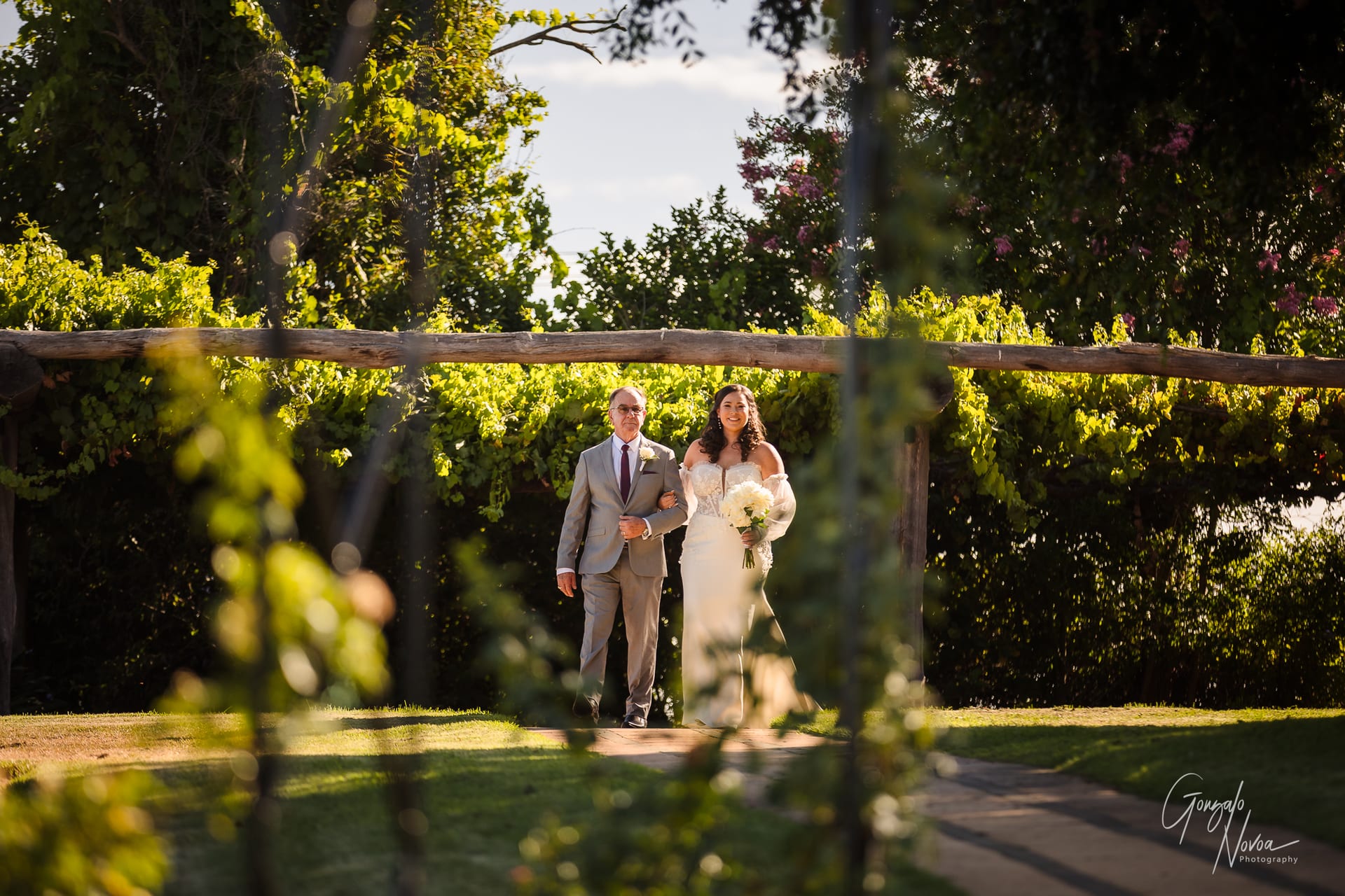 Bride in white dress walks arm-in-arm with an elderly man in a suit under a wooden archway with greenery. Sunlight filters through trees.