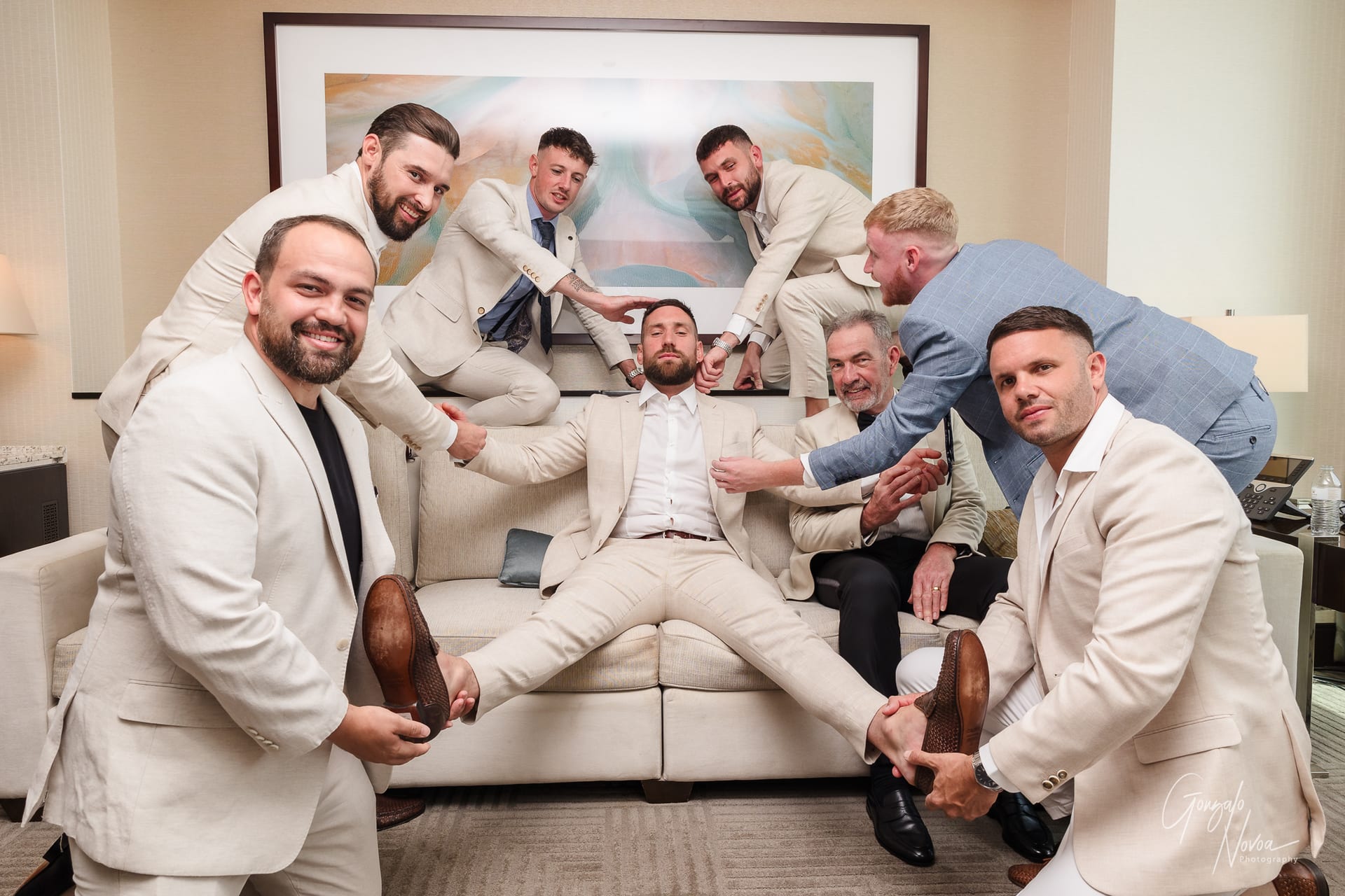 Groomsmen in beige suits playfully pose around the groom on a couch in a hotel room, one man in blue. Cheerful mood, abstract painting behind.