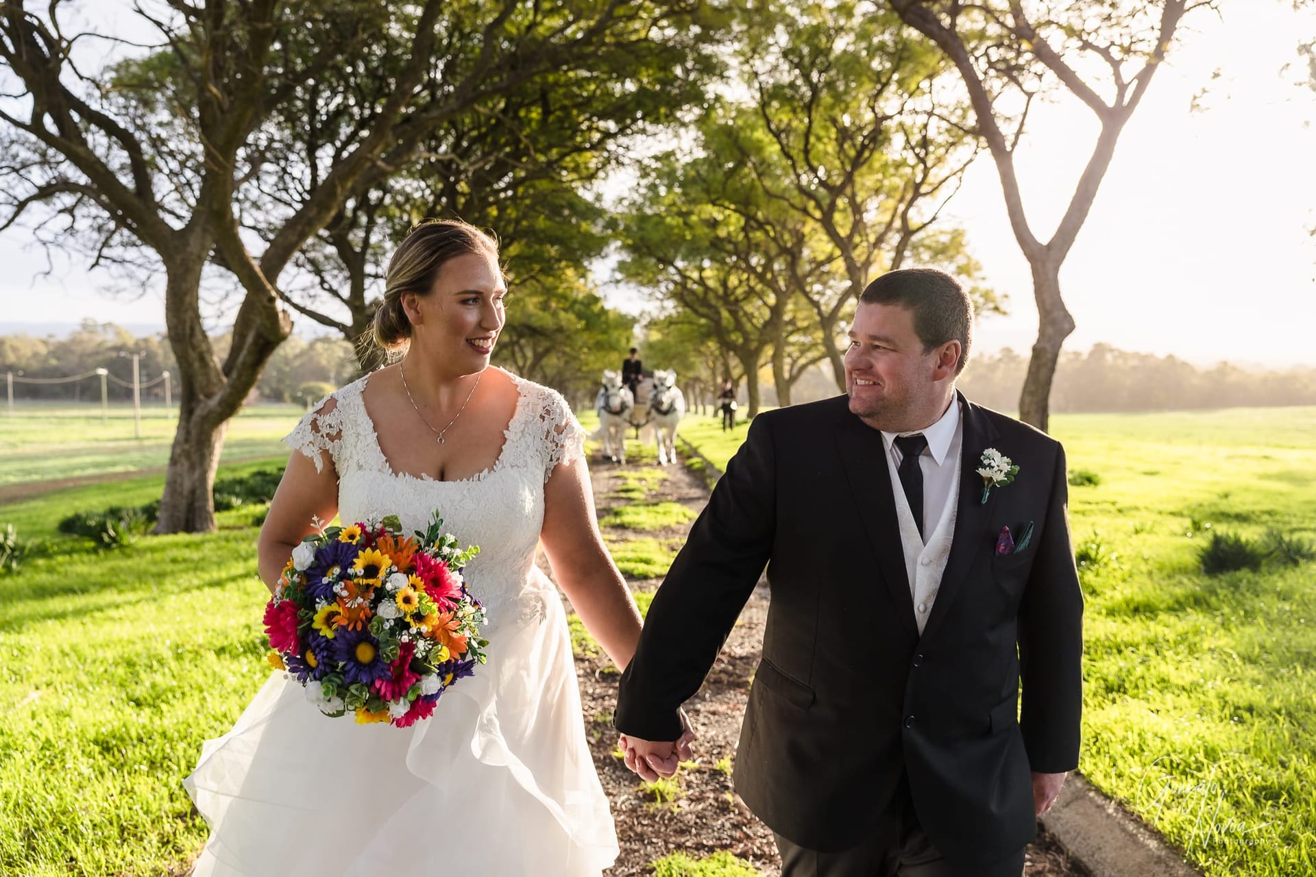 Sarah and James walk hand-in-hand down a tree-lined path at Quarry Farm, smiling at each other as golden light fills the scene, with a white horse-drawn carriage in the background.