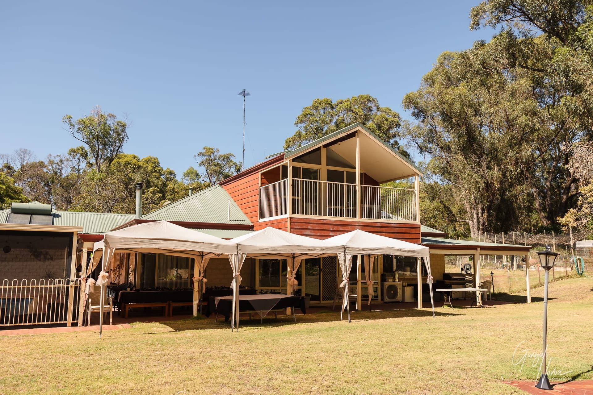 A charming private property with a green roof and wooden balcony, set against a backdrop of tall trees and a clear blue sky. White marquees are arranged on the lawn, creating a welcoming space for Karissa & Josh’s intimate wedding celebration.