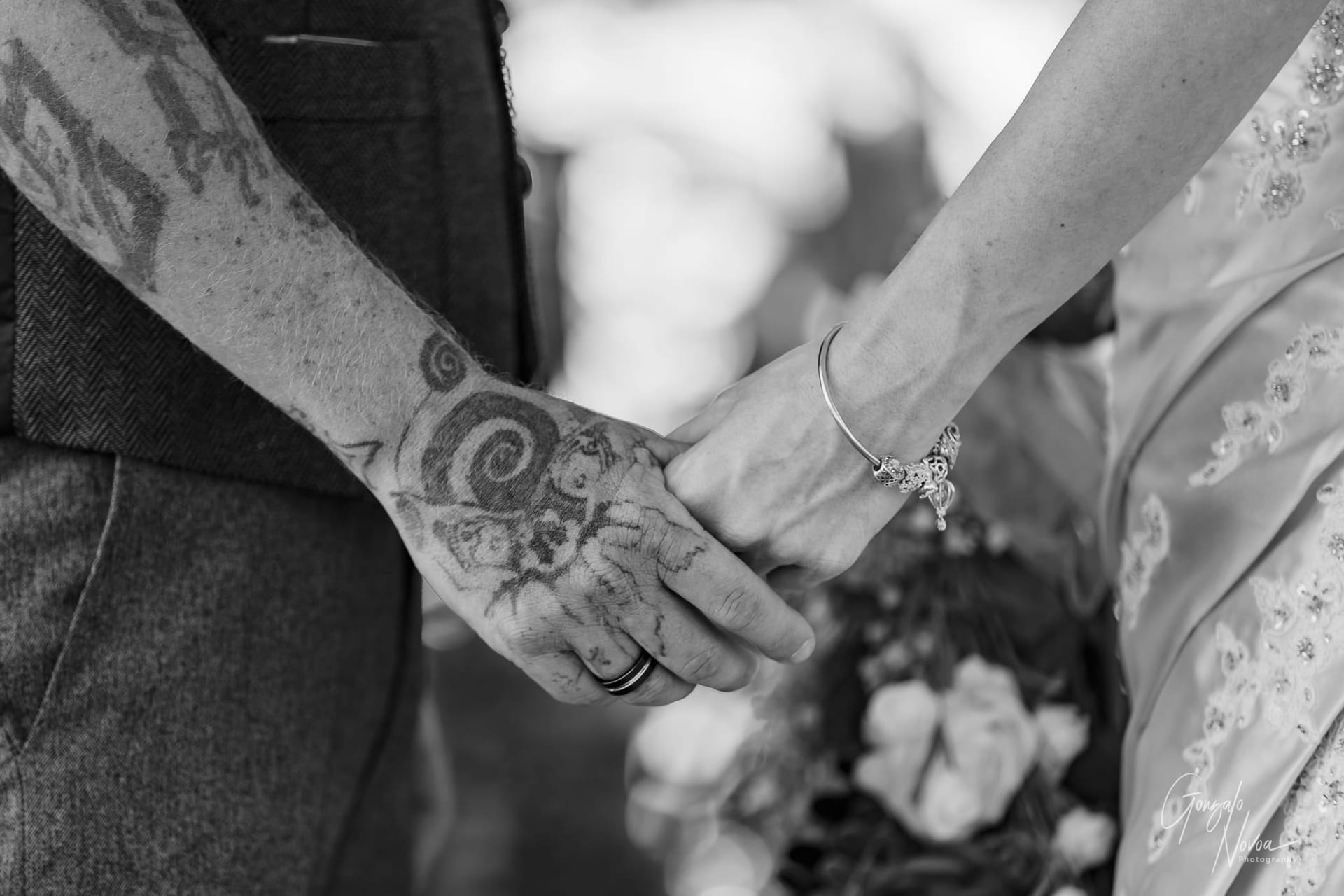 A close-up of Karissa and Josh holding hands, showcasing their wedding rings. Josh’s hand features intricate tattoos, while Karissa wears a delicate rose gold bracelet. A bouquet of vibrant flowers is softly blurred in the background, adding a romantic touch to the intimate moment.