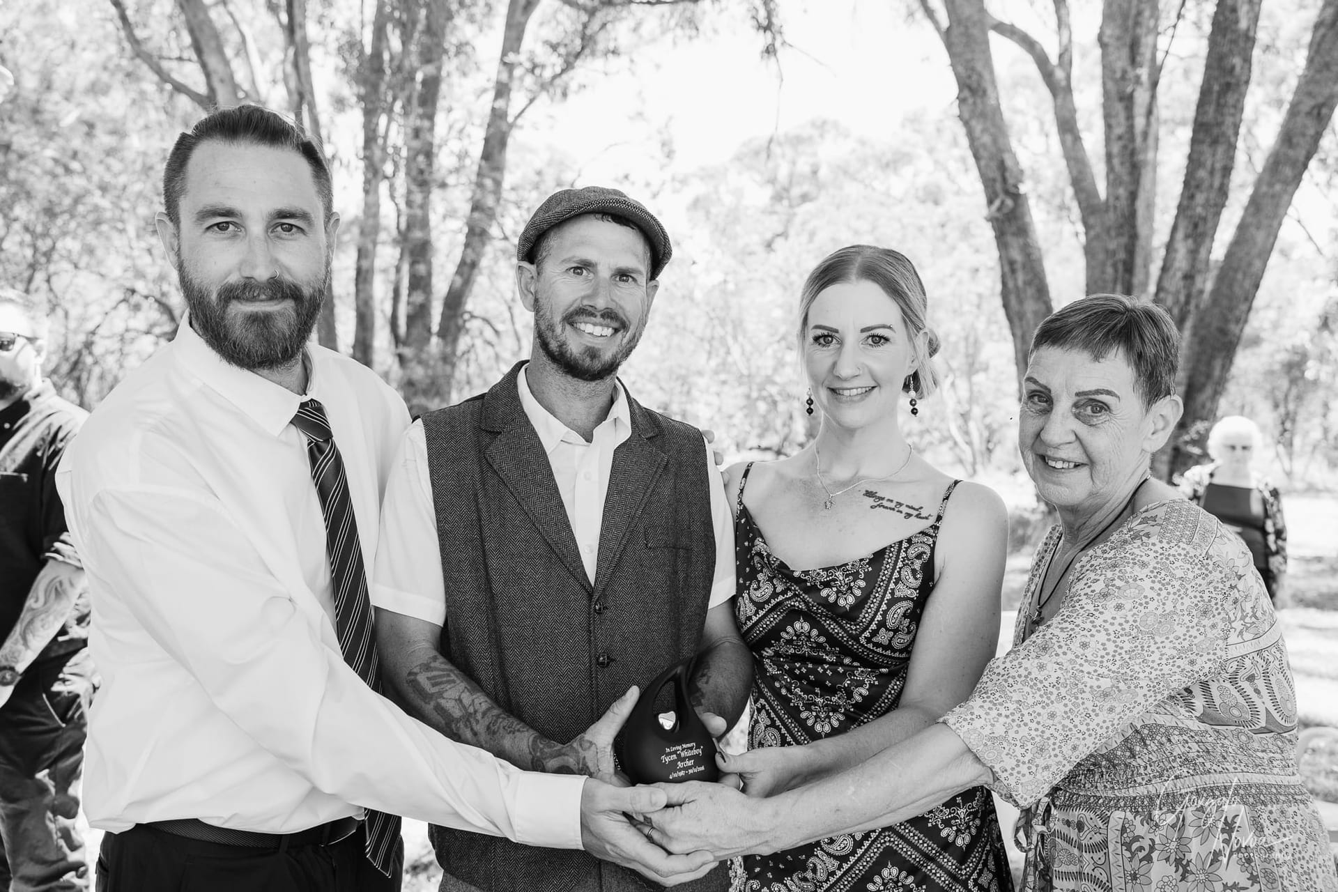 A group of four family members, including the groom, stand together outdoors, smiling while holding a black urn with gold lettering. Sunlight filters through the trees, creating a warm and heartfelt moment of remembrance during the wedding celebration.