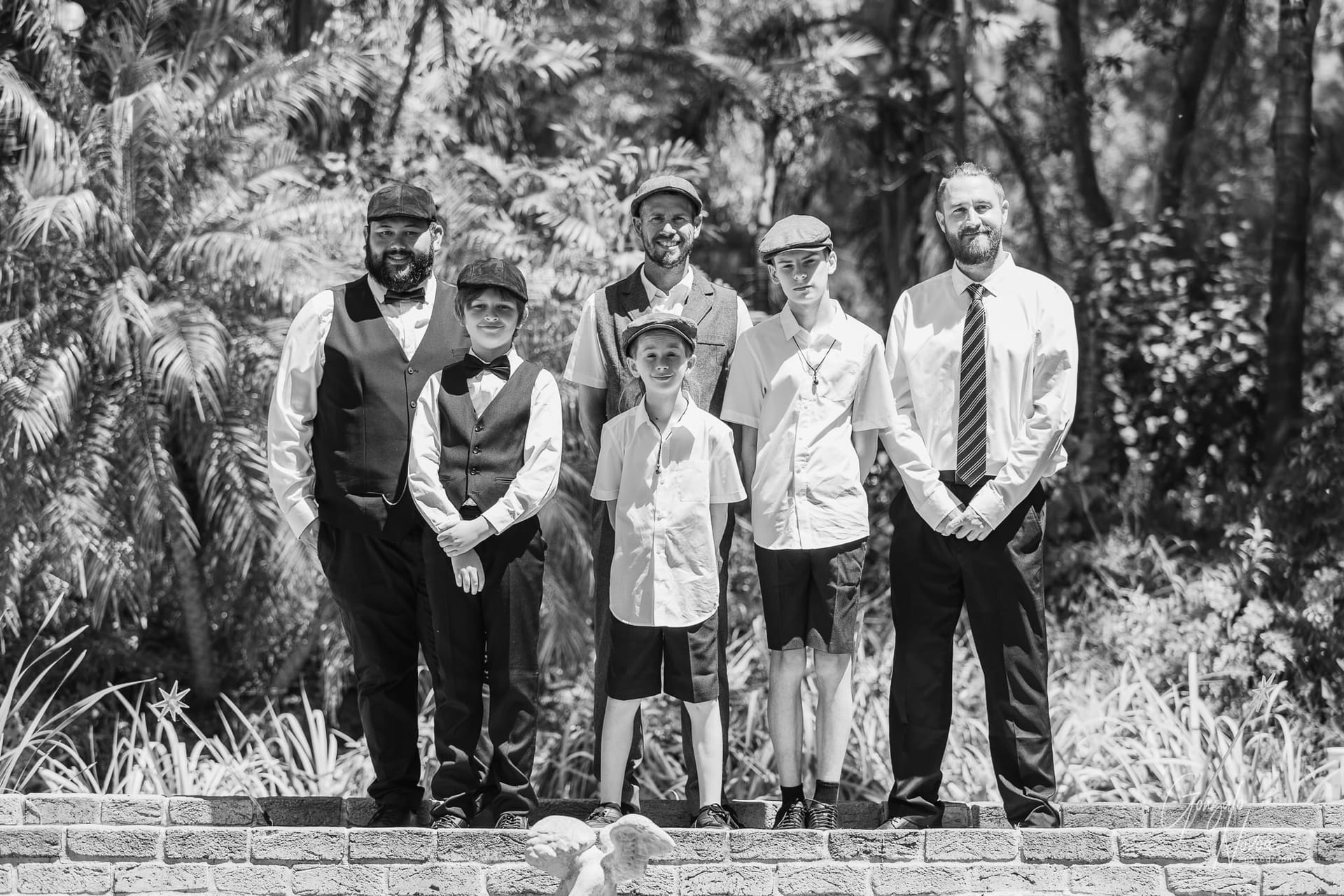 A group portrait of Josh standing alongside his two sons and family members, dressed in a mix of formal and vintage-inspired attire. The group poses on a brick ledge with lush greenery behind them, capturing a meaningful family moment on the wedding day.