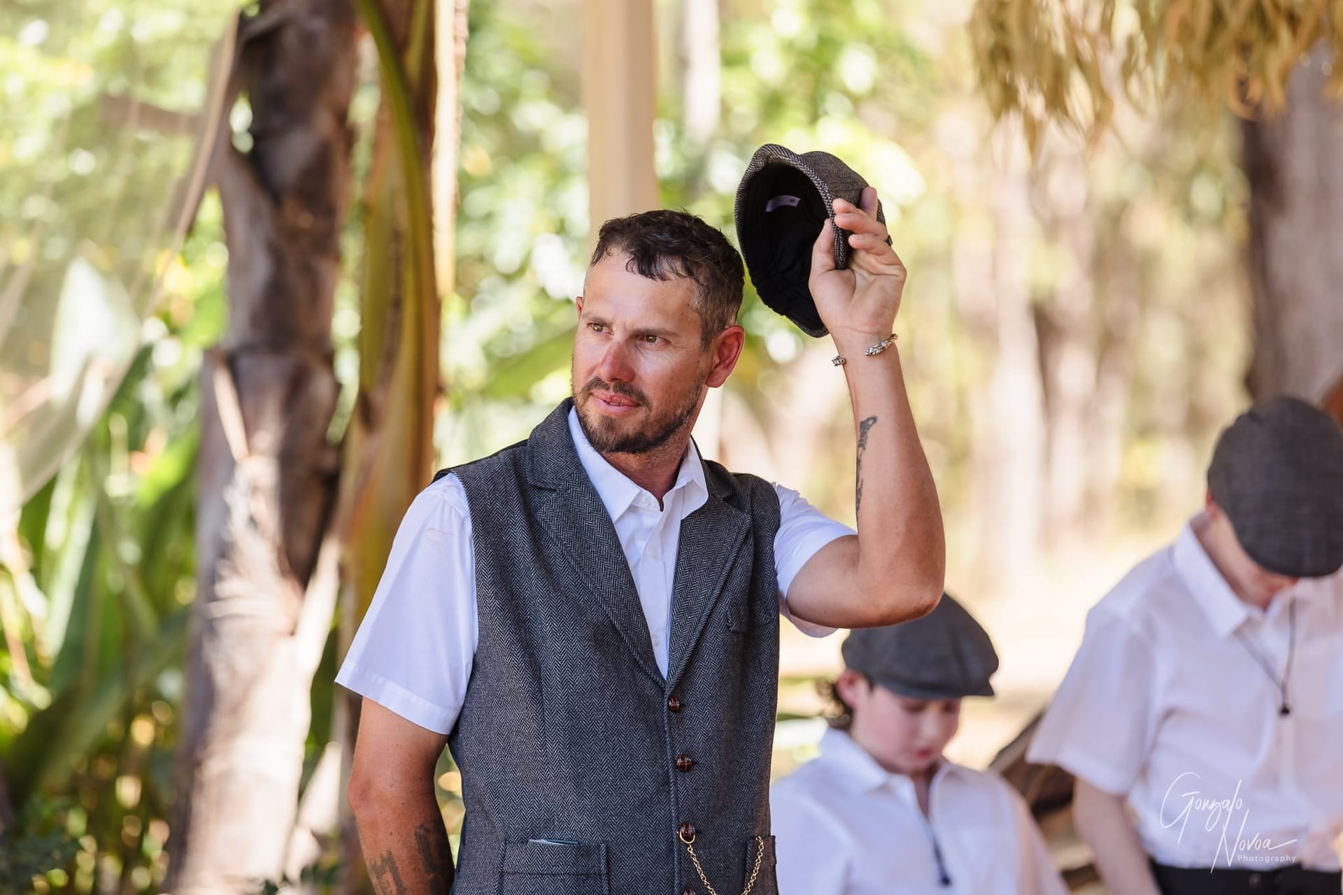 Josh in a gray vest holds up a flat cap outdoors, with greenery in the background. The kids in similar attire are behind him.