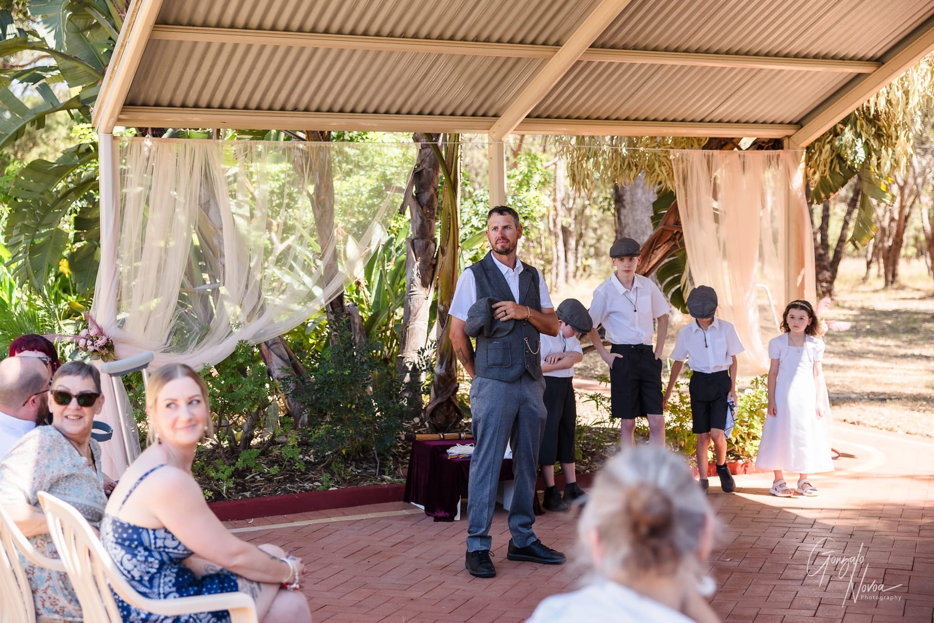 Josh in a vest stands under a pavilion with his children in caps and shorts. Guests seated nearby. Trees and draped fabric in the background.