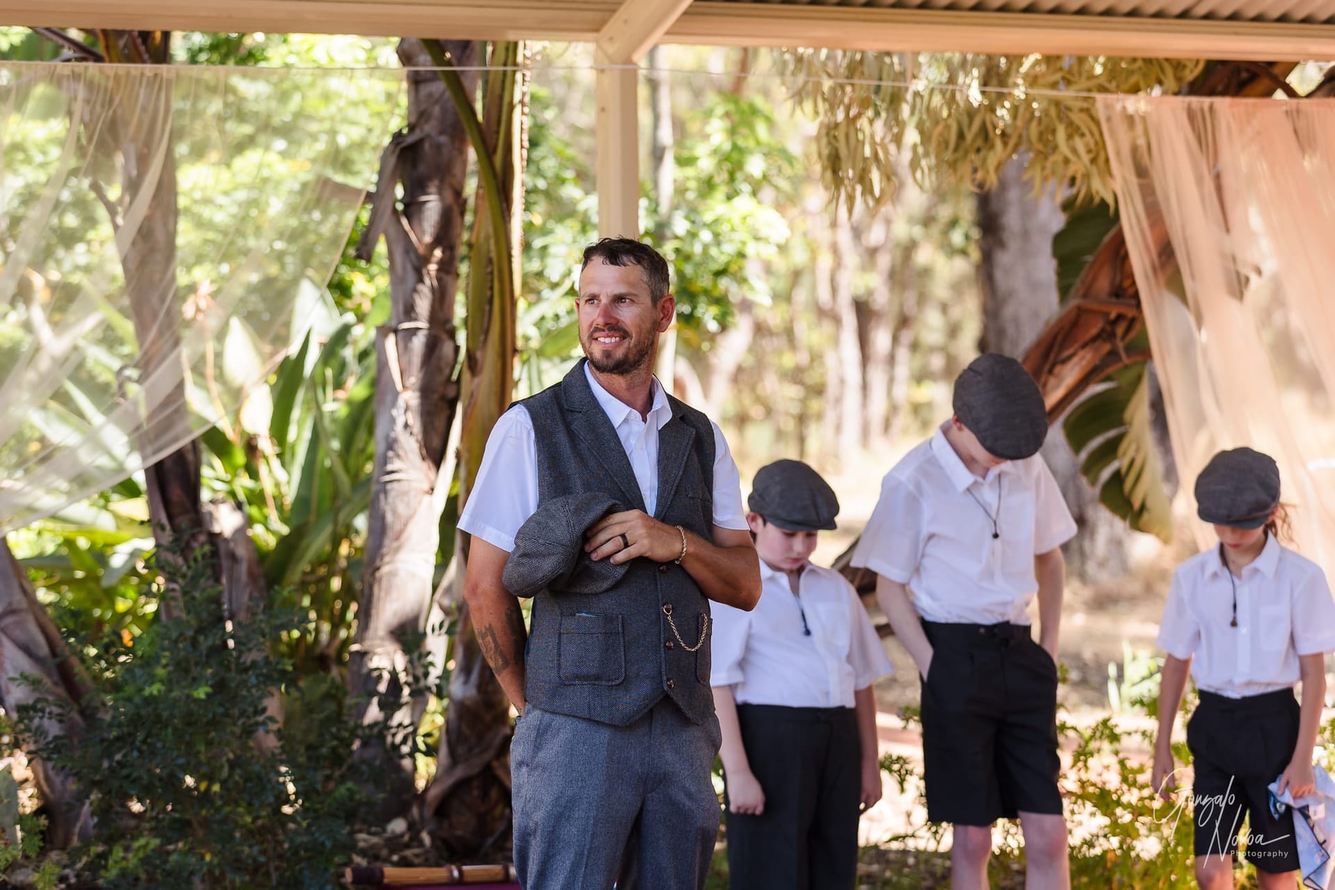Josh in grey vest holding cap, standing outdoors with his three boys in white shirts and caps. Greenery and trees in the background. Relaxed mood.