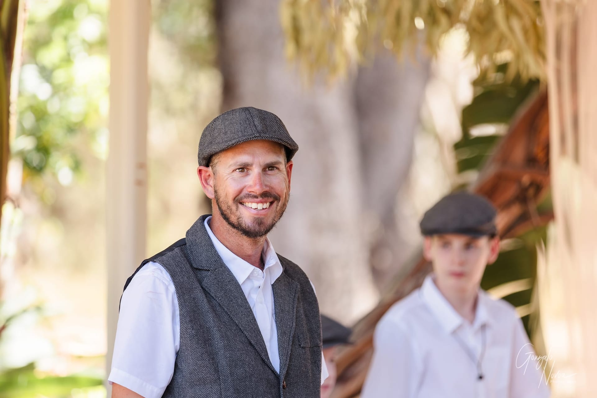 Josh smiling in a gray vest and flat cap, with one of his sonds behind. Background is outdoors with green foliage and soft lighting.