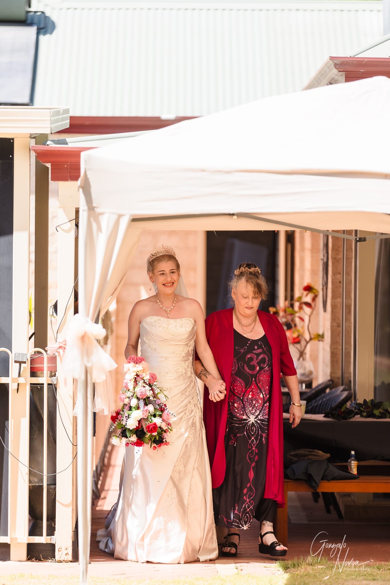 Karissa with her gown and tiara with a bouquet walks hand in hand with a Janine in a red outfit under a white canopy, smiling.