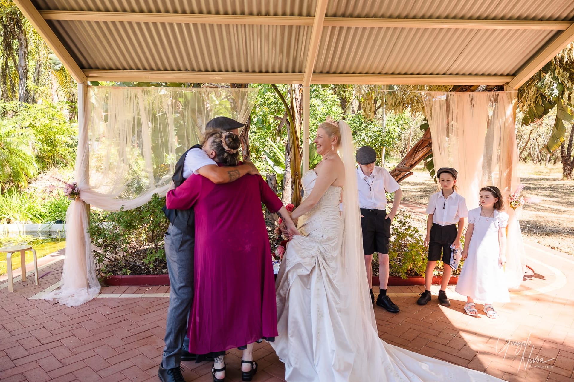 Under a beautifully decorated outdoor pavilion, Josh embraces Janine, Karissa’s mum, in a heartfelt hug while holding hands with Karissa, who smiles warmly in her wedding gown. Nearby, their children, dressed in vintage-style white shirts and suspenders, watch the moment unfold. The setting is surrounded by lush greenery, with soft drapery adding a romantic touch to the scene.