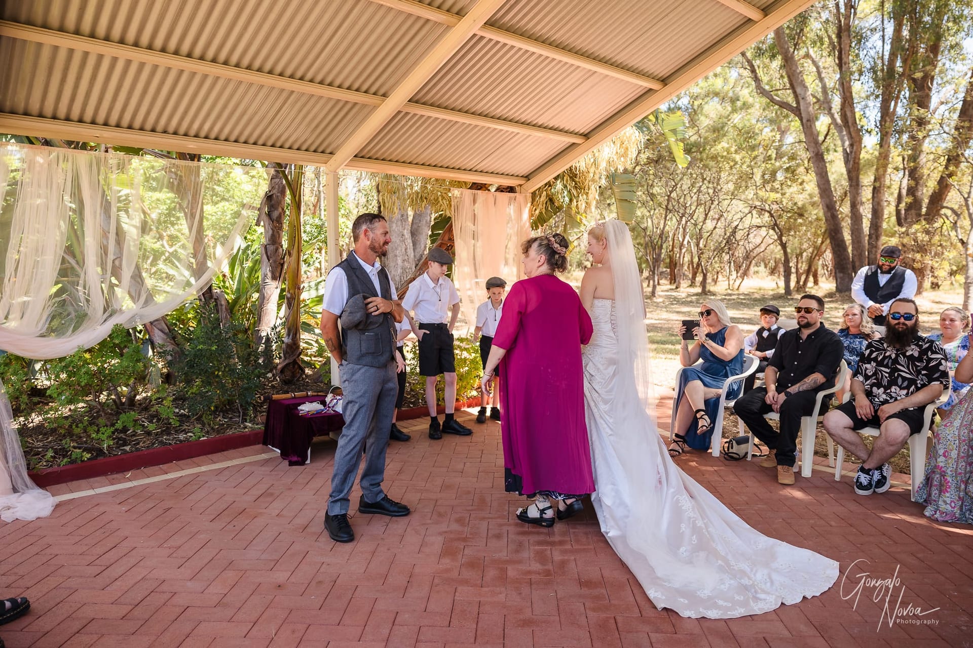 Wedding ceremony under a gazebo. Karissa in her wedding dress with veil. Guests seated around, trees in the background, sunny day.