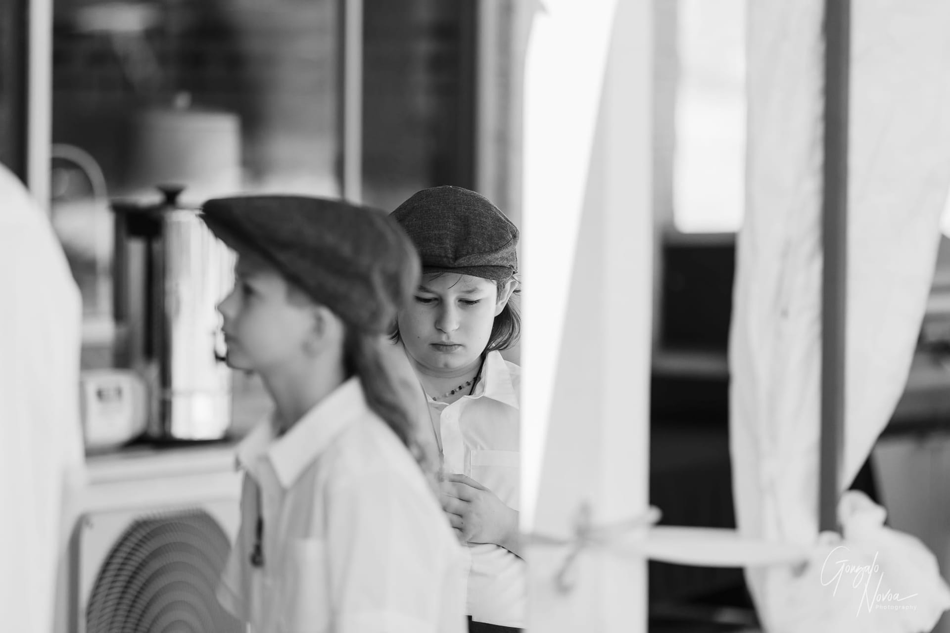 A young child dressed in a white shirt and vintage-style flat cap stands in quiet reflection, looking down with a thoughtful expression. Another child in similar attire is slightly out of focus in the foreground, capturing the anticipation and emotion of the wedding day.