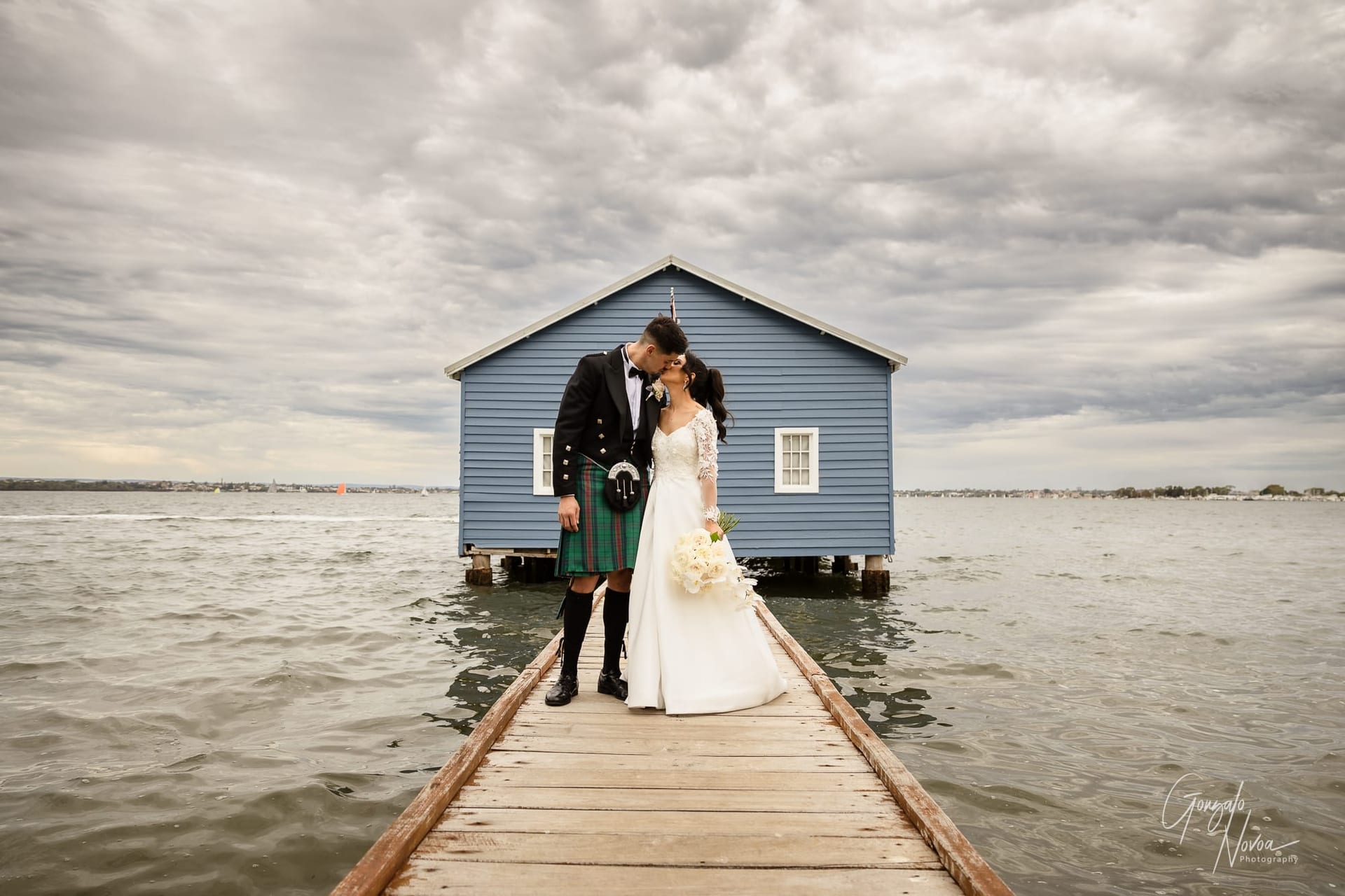 Bride and groom share a kiss on a wooden pier by a blue boathouse. Overcast sky, groom in kilt, bride holds bouquet. Romantic mood.