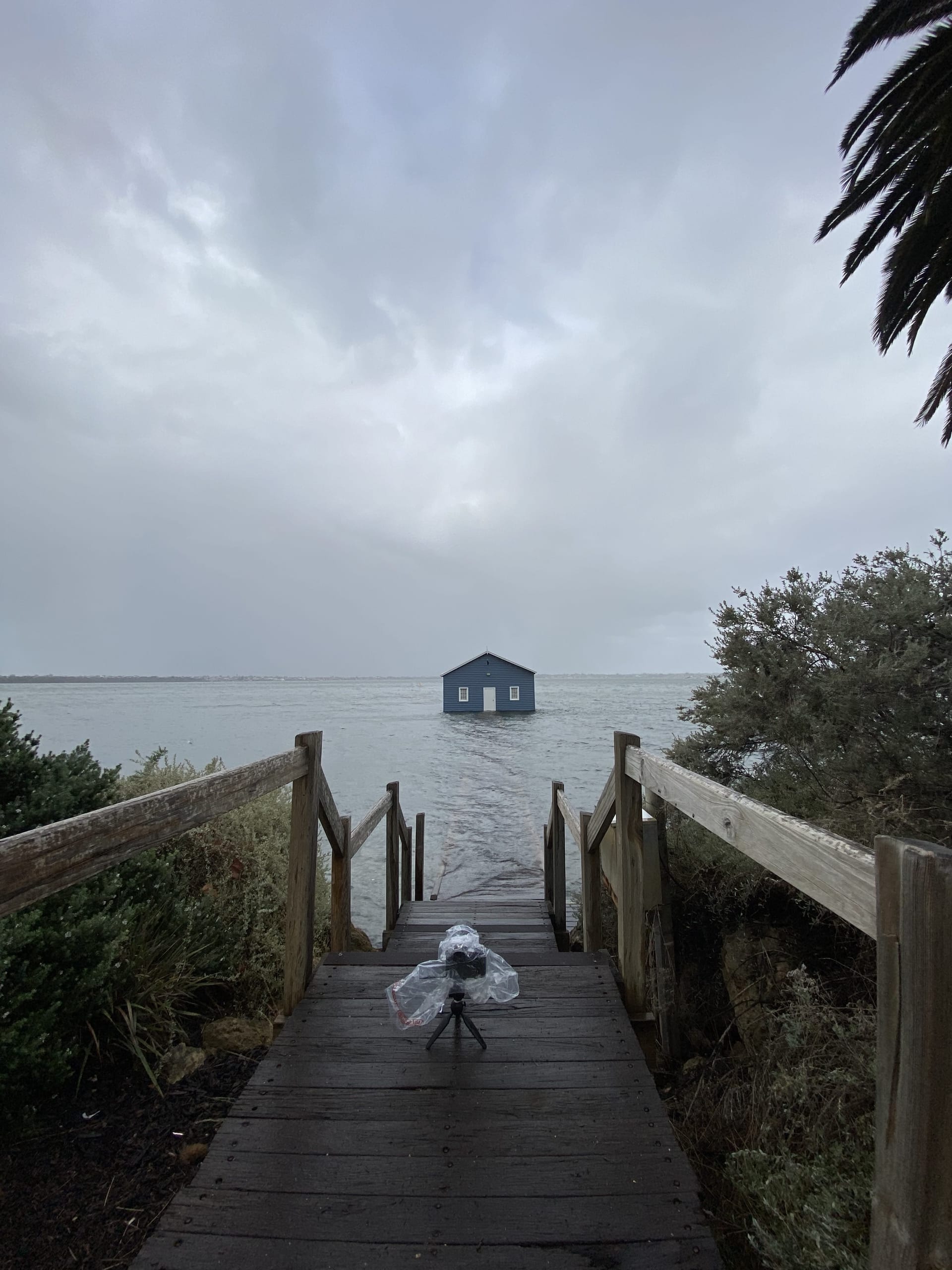 A camera on a tripod covered in a plastic bag sits on a wooden walkway leading to Perth’s Blue Boat House, with stormy skies and choppy waters in the background.
