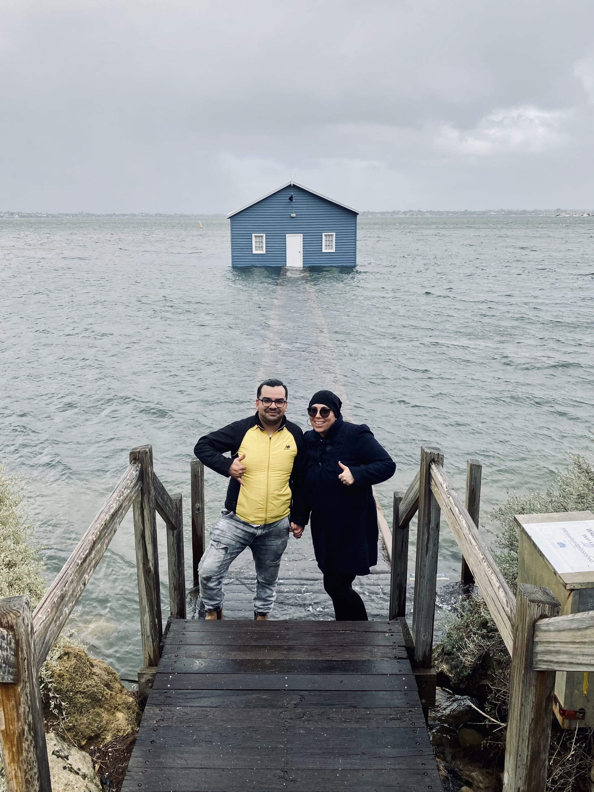 A couple standing on the wooden walkway in front of the Blue Boat House in Perth, dressed in wet-weather gear, smiling and giving thumbs up despite the stormy conditions.