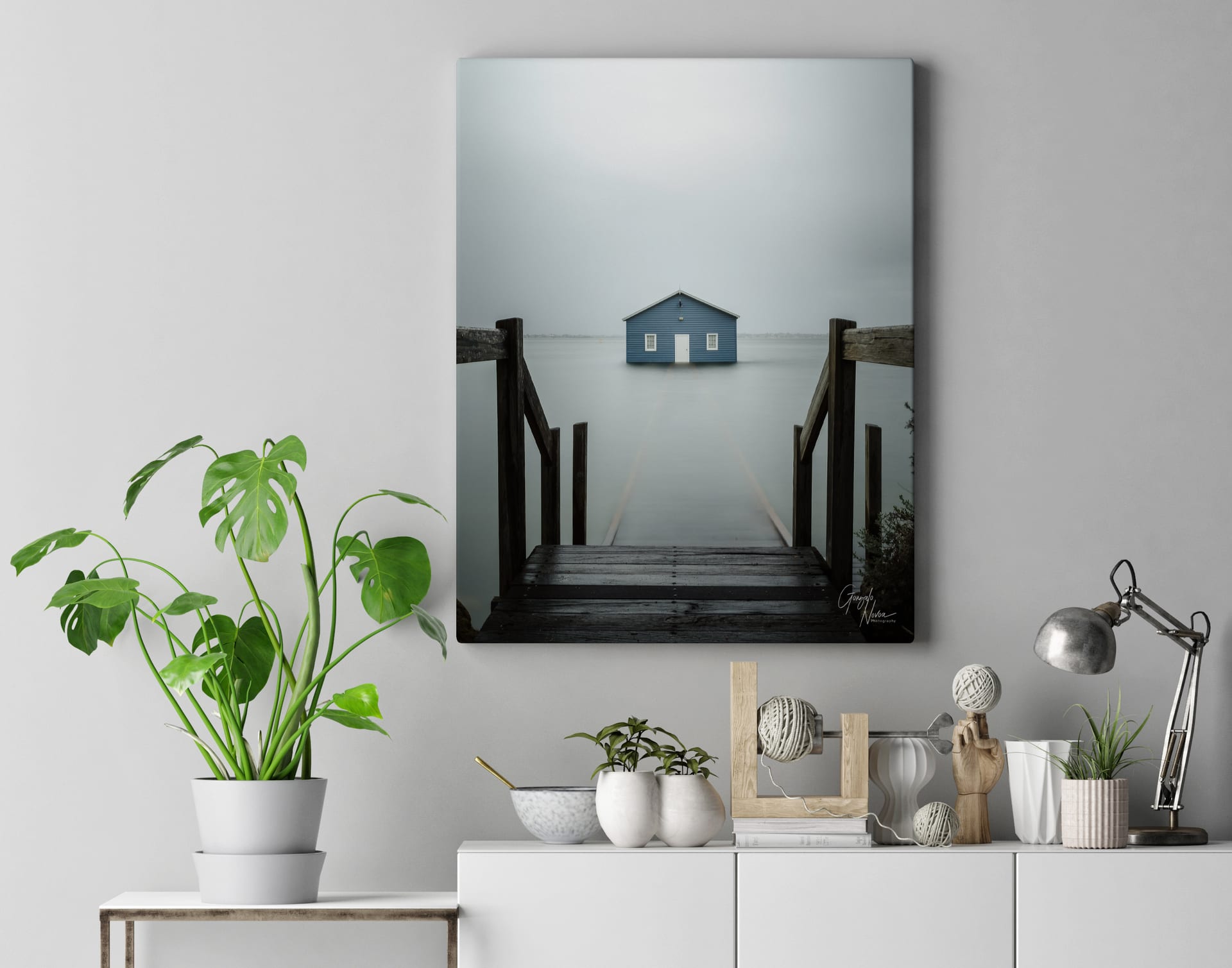 A modern interior featuring a large canvas print of the Blue Boat House, showcasing a misty and minimalist aesthetic. The print is displayed on a white wall above a stylish sideboard with indoor plants and decorative elements.