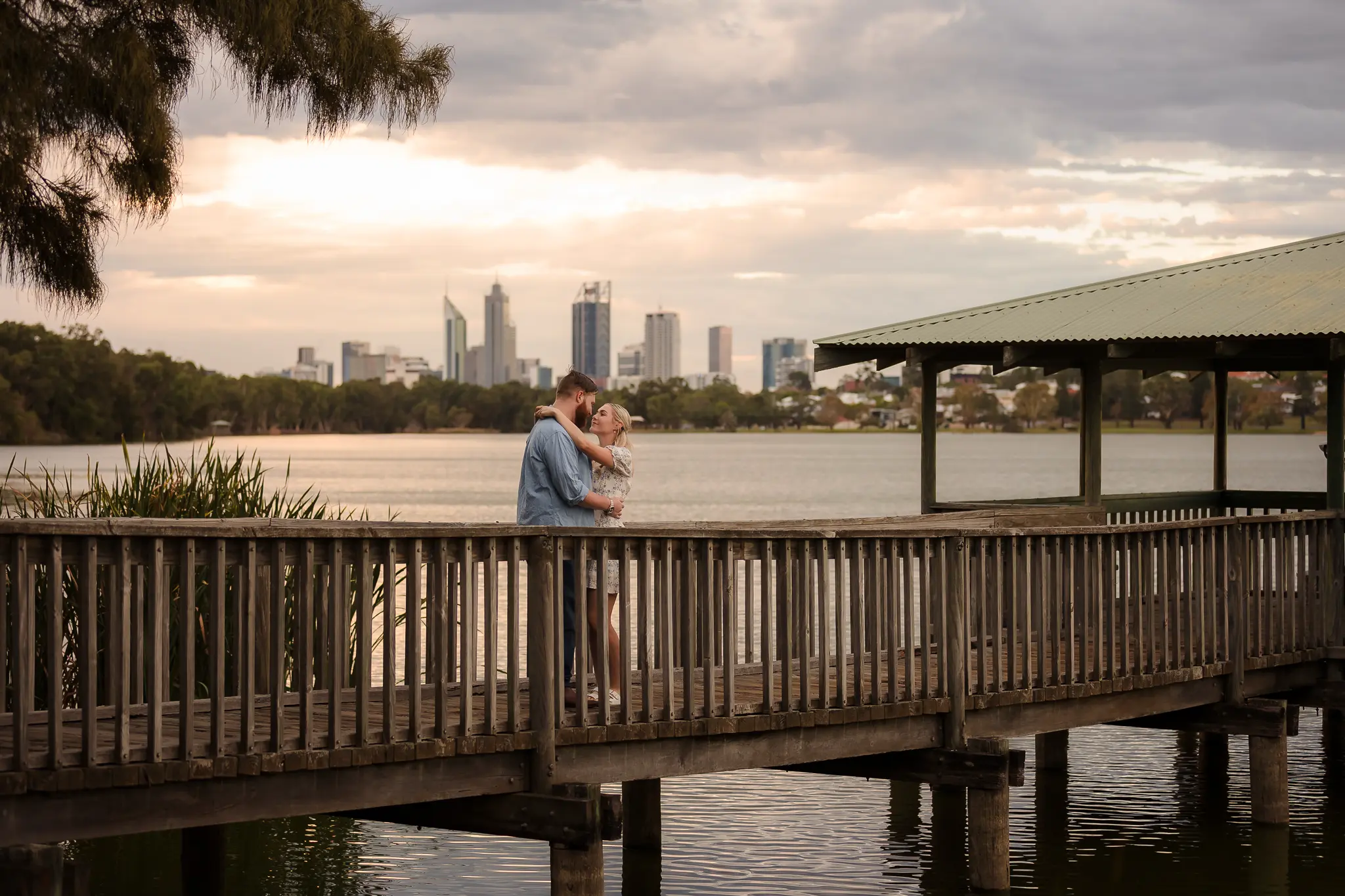 Couple embracing on a wooden pier at sunset. City skyline in the distance, calm water below, creating a romantic and serene mood.