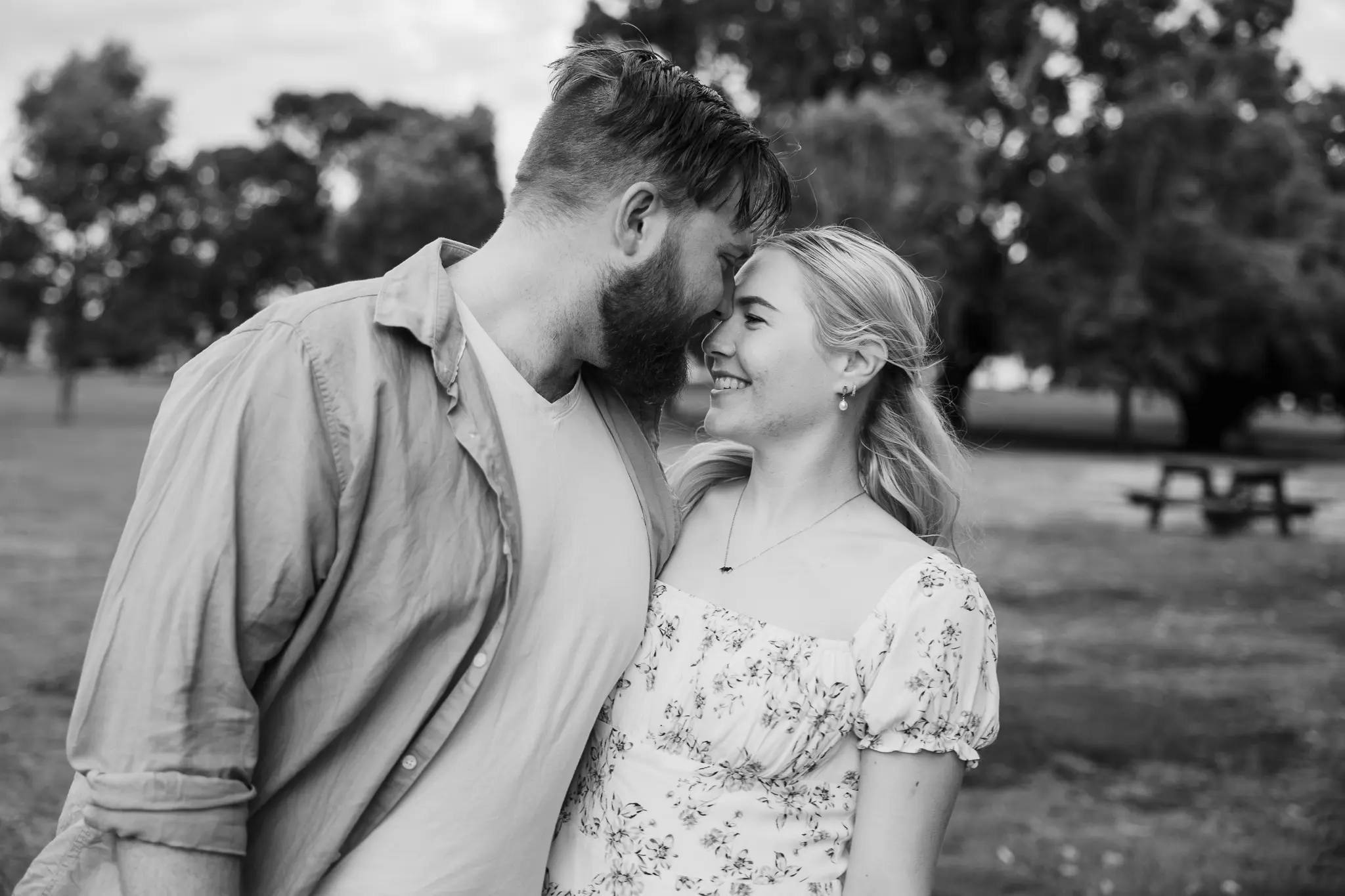 A couple smiles affectionately forehead to forehead in a park. The woman wears a floral dress. Trees and a picnic table are in the background.